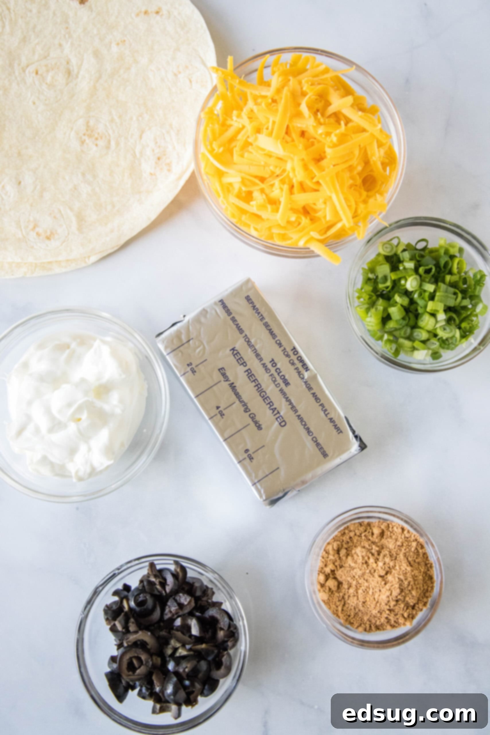 Various ingredients for taco pinwheels laid out on a white surface, including cream cheese, sour cream, taco seasoning, cheese, olives, and green onions.