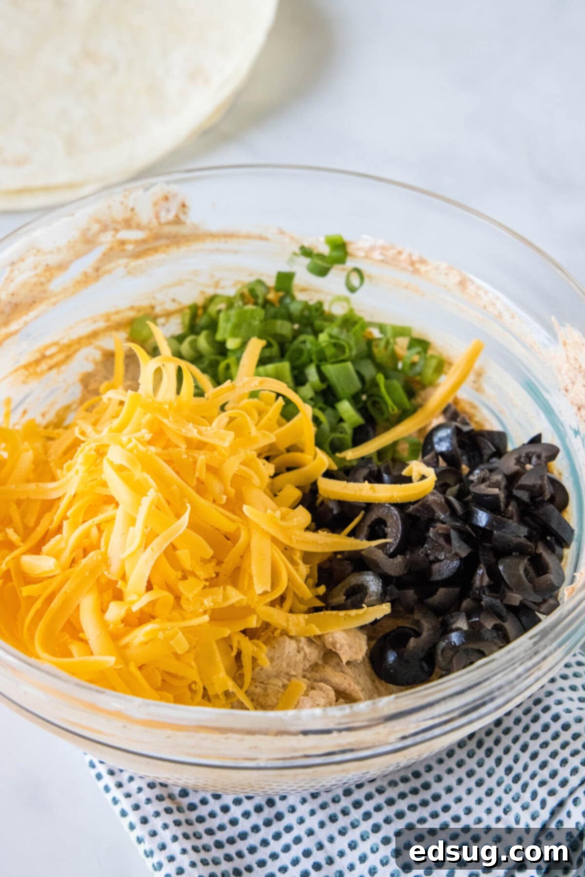 Shredded cheese, black olives, and chopped green onions are carefully folded into the seasoned cream cheese filling in a glass bowl.