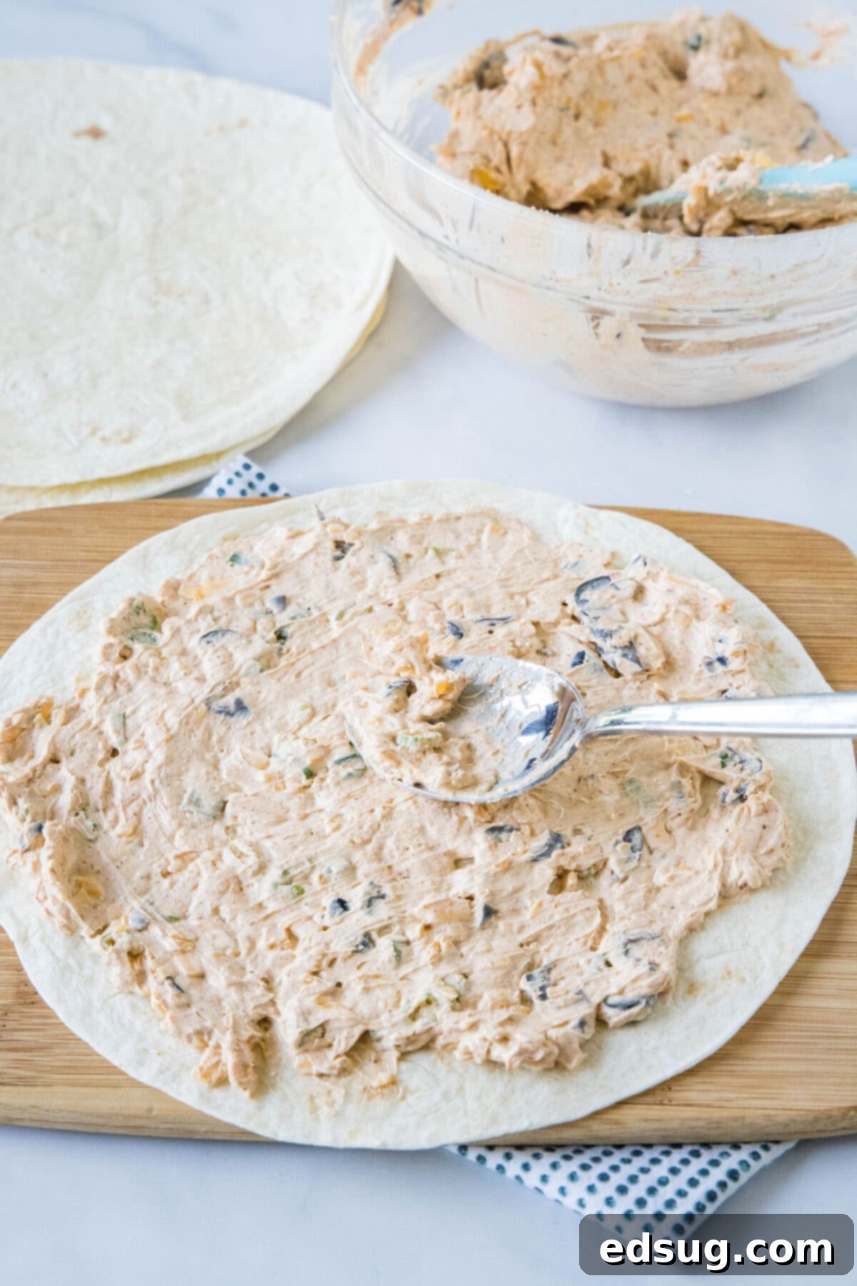 A spoon is used to smoothly spread the prepared taco cream cheese filling evenly over a soft flour tortilla, with more filling and tortillas visible in the background.