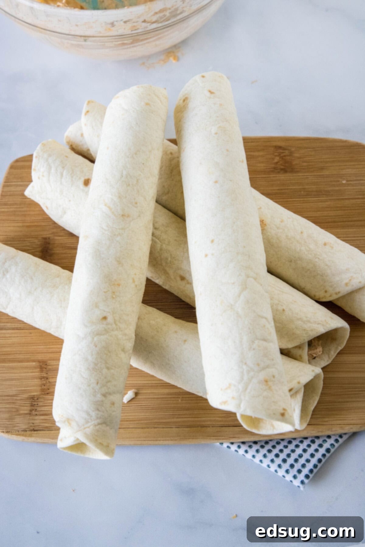 An overhead view showing several tightly rolled flour tortillas, filled with the creamy taco mixture, neatly stacked on a wooden cutting board, awaiting refrigeration.