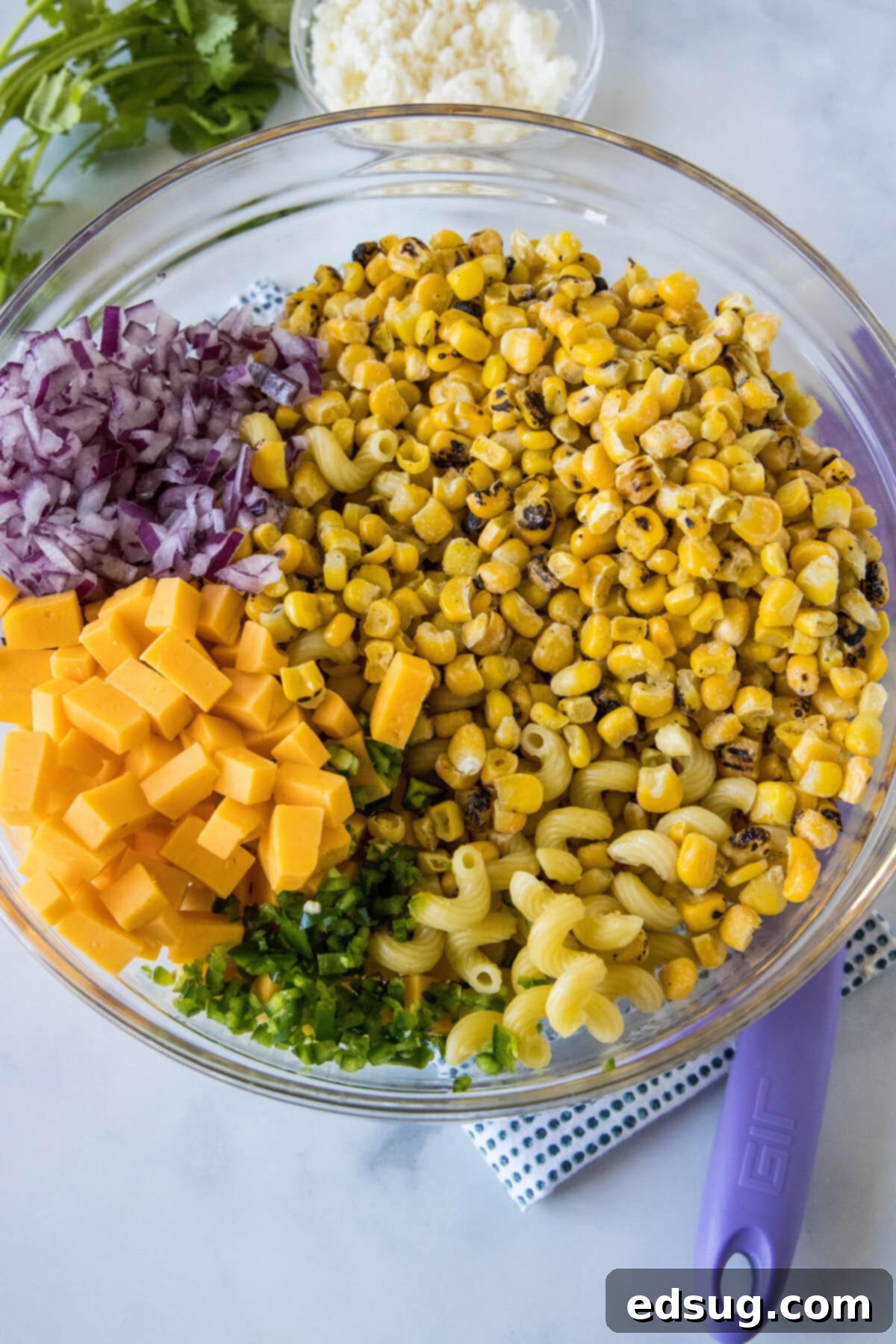 All the vibrant ingredients for street corn pasta salad, including cooked pasta, roasted corn, and fresh vegetables, gathered in a large glass bowl.