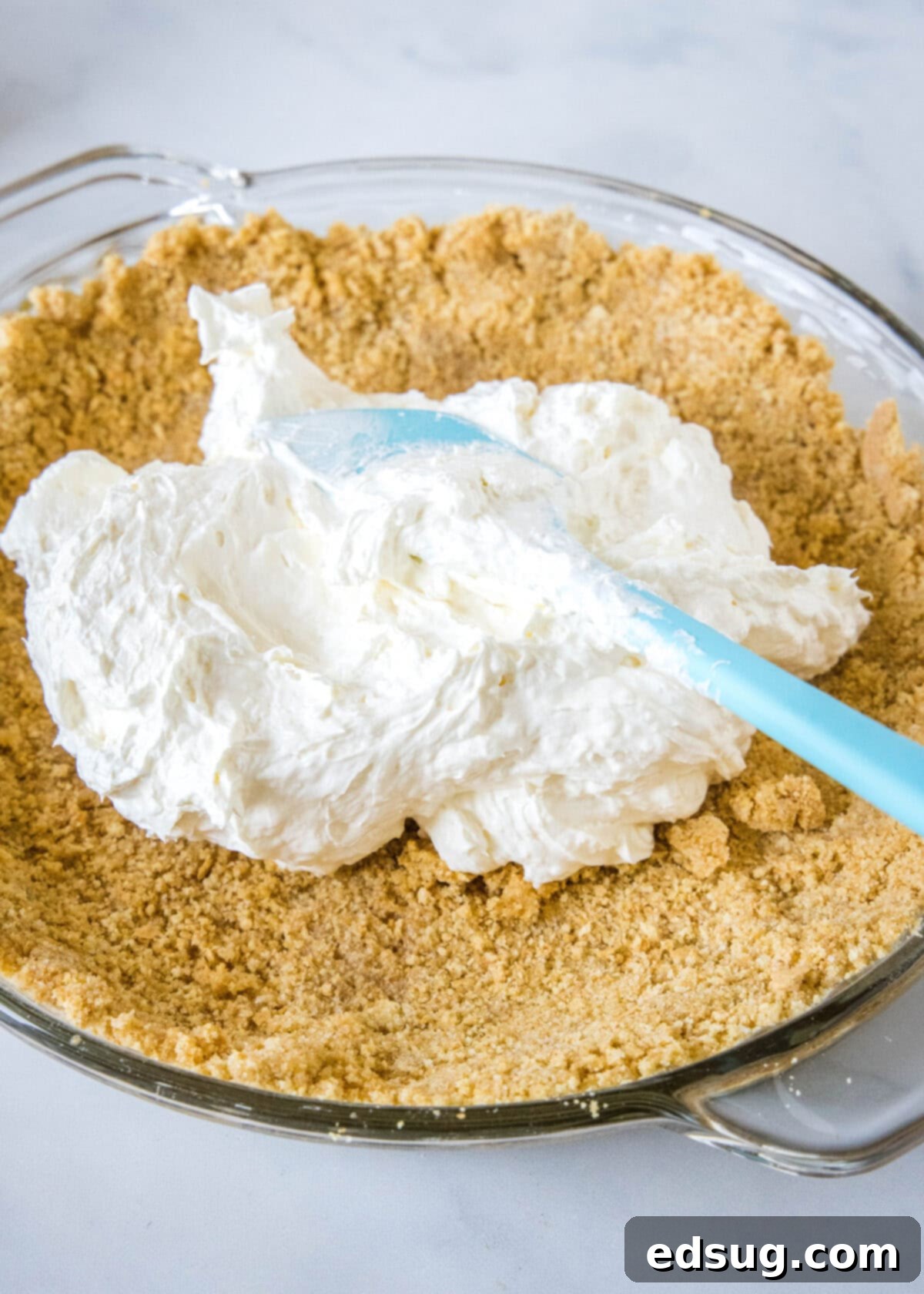 Banana split pie is a cool and creamy no-bake dessert that you can make in 20 minutes. Filled with whipped cream, bananas, and pineapple! Smooth, creamy cream cheese pie filling being spread evenly into a golden graham cracker crust using a blue spatula, preparing the base for the pie layers.