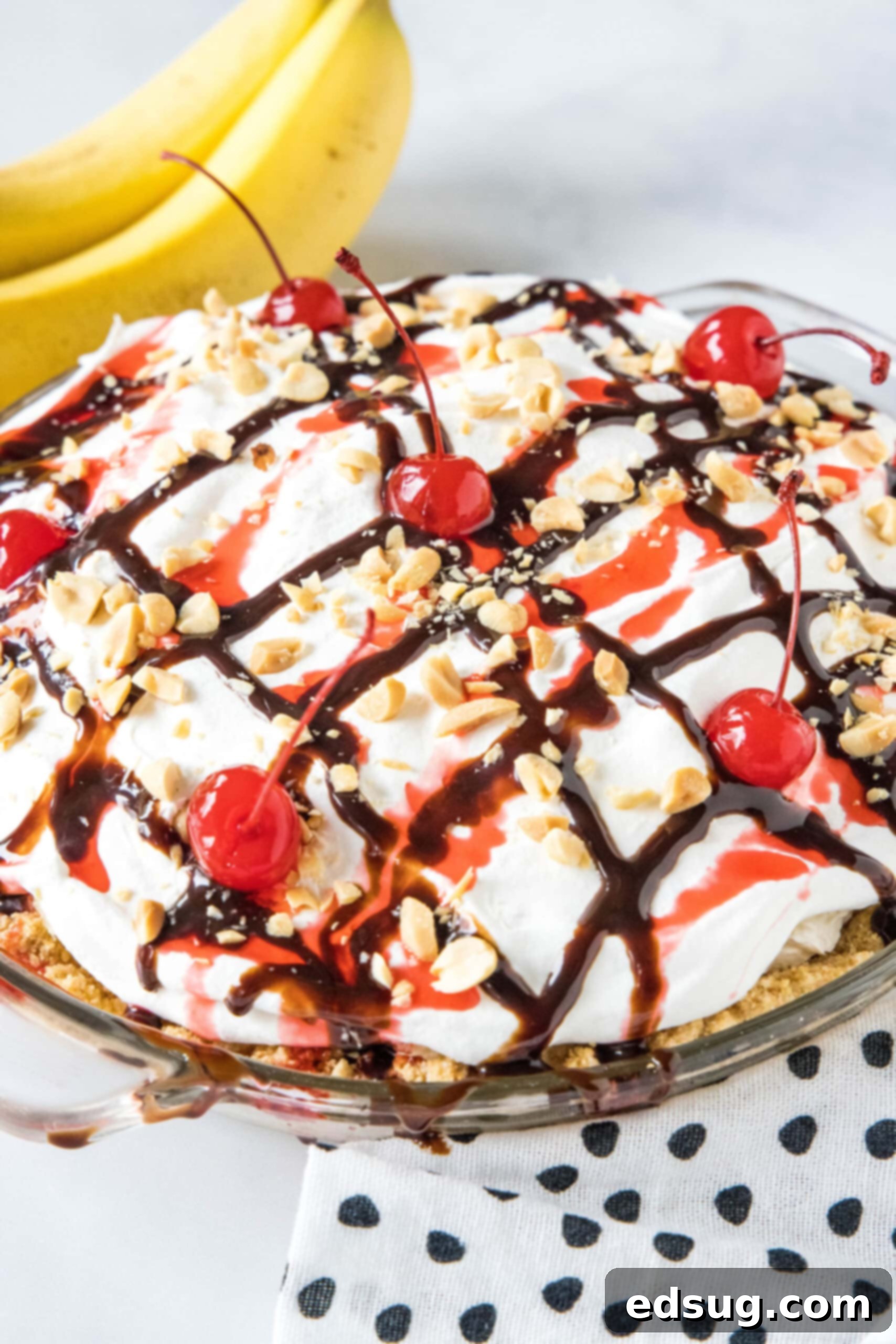 Banana split pie is a cool and creamy no-bake dessert that you can make in 20 minutes. Filled with whipped cream, bananas, and pineapple! A stunning full view of a completed banana split pie, beautifully drizzled with rich chocolate and sweet strawberry sauces, topped with crunchy chopped peanuts and bright red cherries, ready to be served.