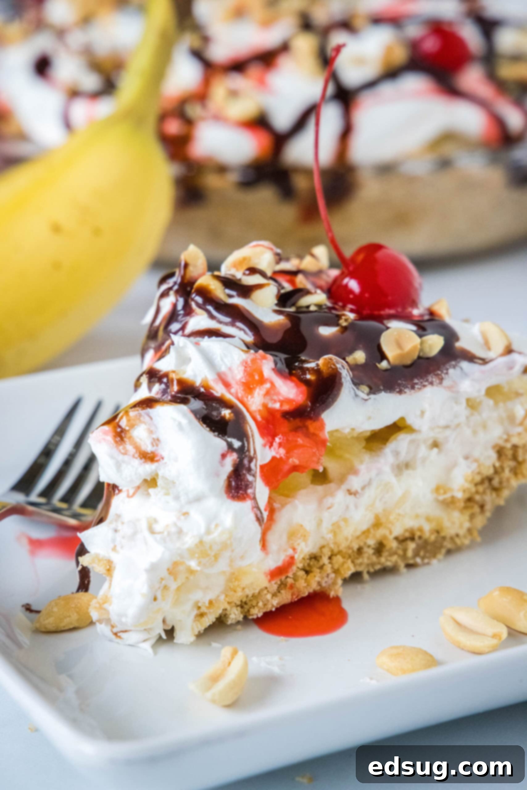 Banana split pie is a cool and creamy no-bake dessert that you can make in 20 minutes. Filled with whipped cream, bananas, and pineapple! A perfectly portioned slice of banana split pie, elegantly arranged on a plate and adorned with chocolate and strawberry drizzles, chopped peanuts, and a cherry, with the rest of the pie and a banana in soft focus in the background.