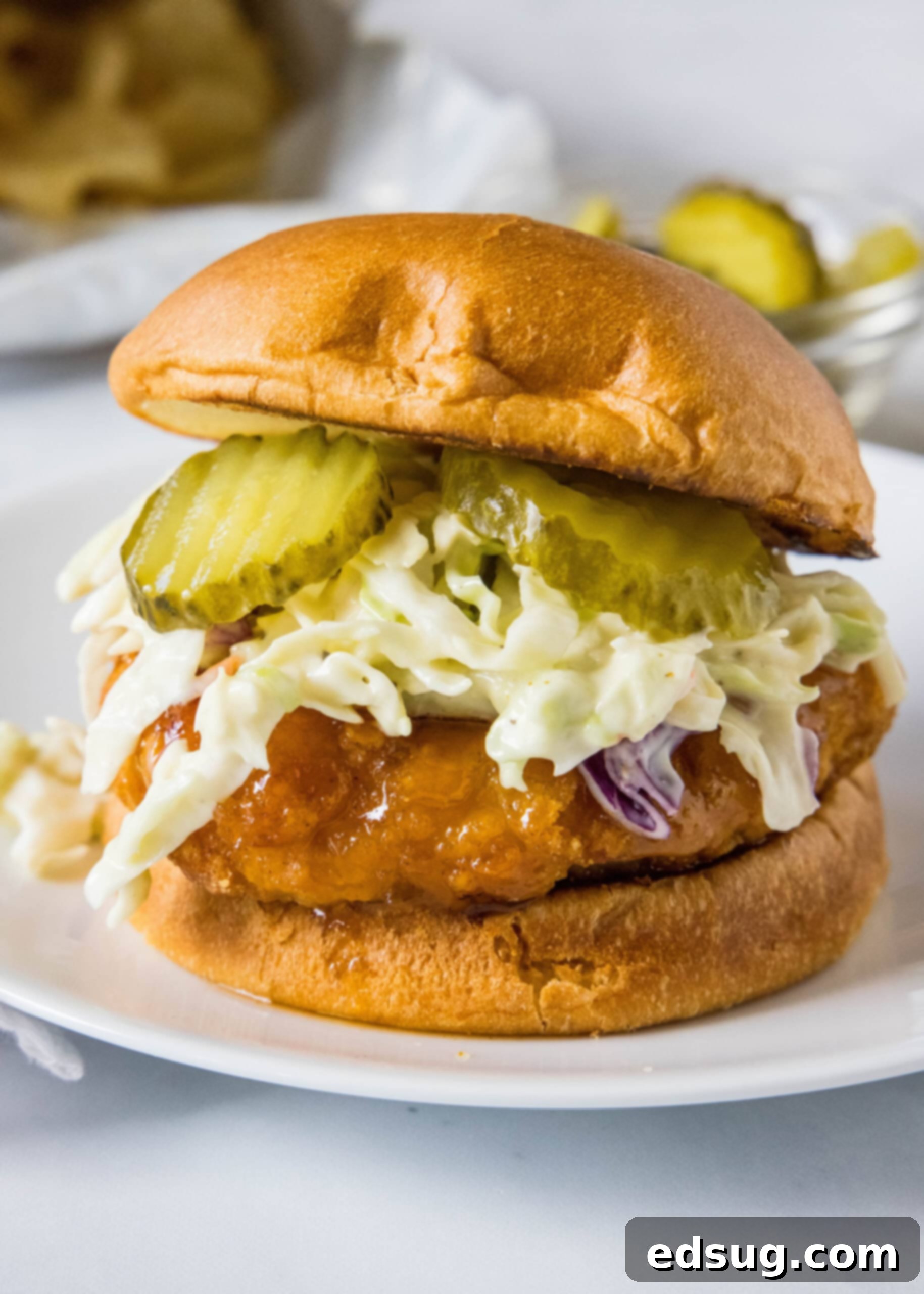 Make a hot honey chicken sandwich with crispy chicken coated in spicy honey, served between fluffy burger buns with cool and crunchy coleslaw! A hot honey chicken sandwich topped with coleslaw and pickles on a white plate.