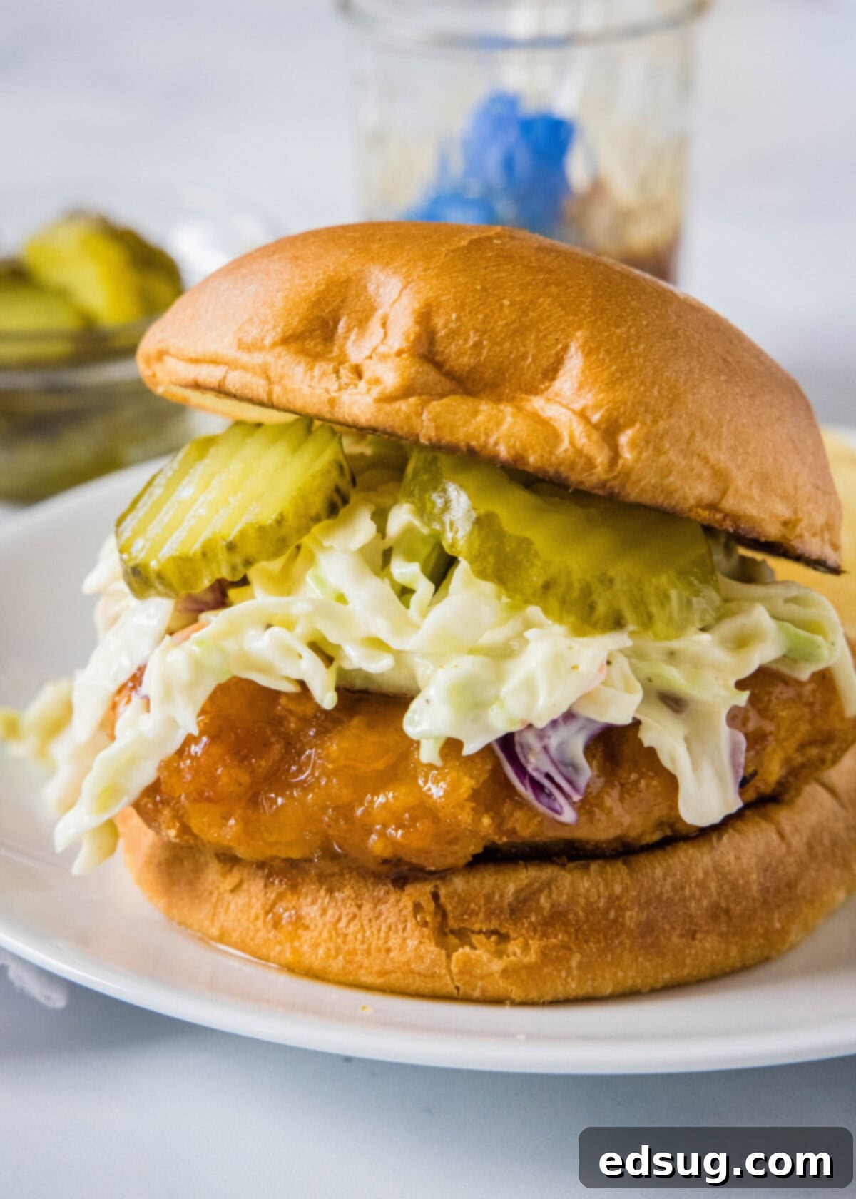 Make a hot honey chicken sandwich with crispy chicken coated with spicy honey, served between fluffy burger buns with cool and crunchy coleslaw! A hot honey chicken sandwich topped with coleslaw and pickles on a white plate.