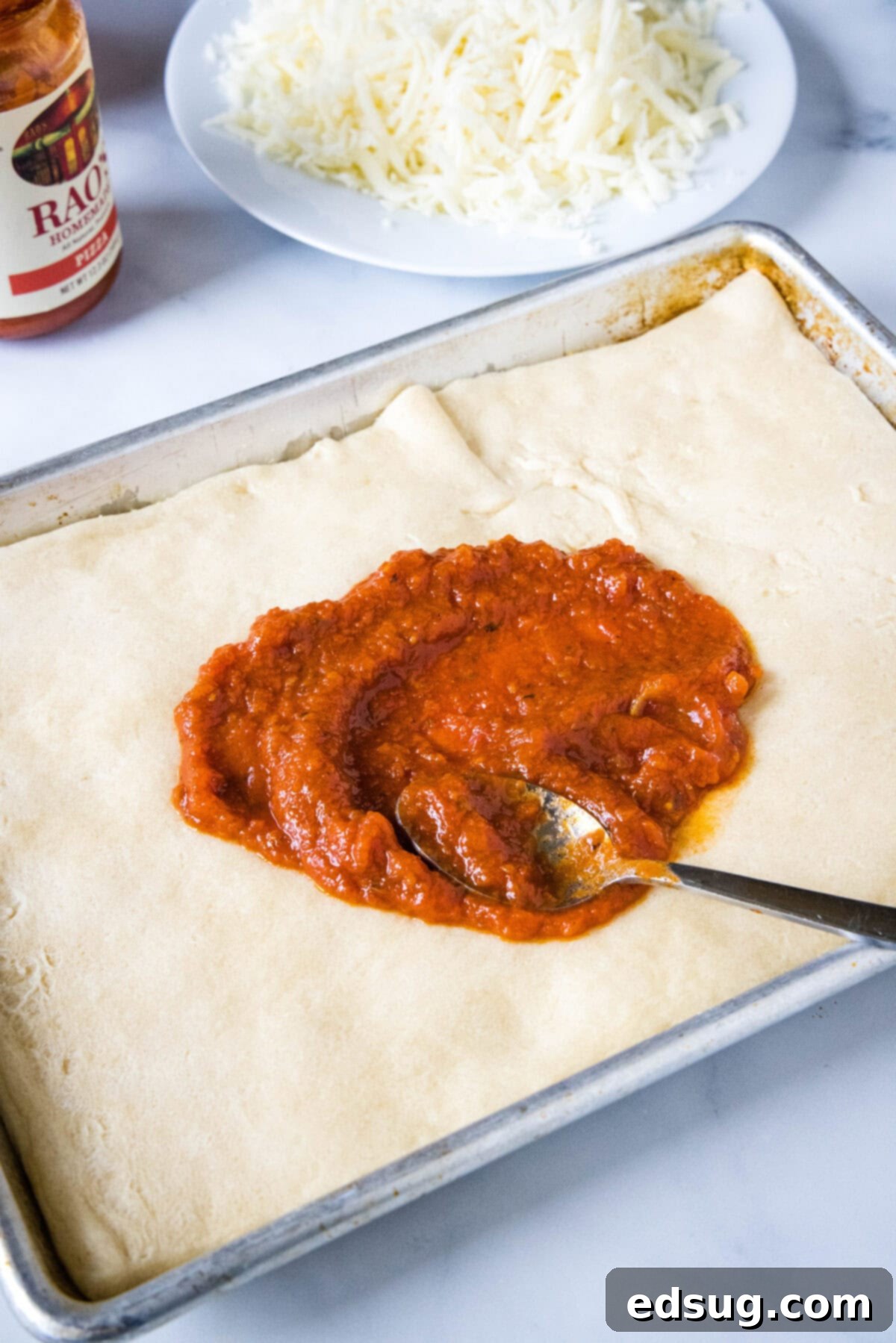 Cheesy crescent roll pizza is the perfect dinner or party snack! There’s zero kneading or rise time, just add your favorite toppings and bake! A spoon resting in a circle of partially spread pizza sauce in the middle of a crescent dough pizza crust.