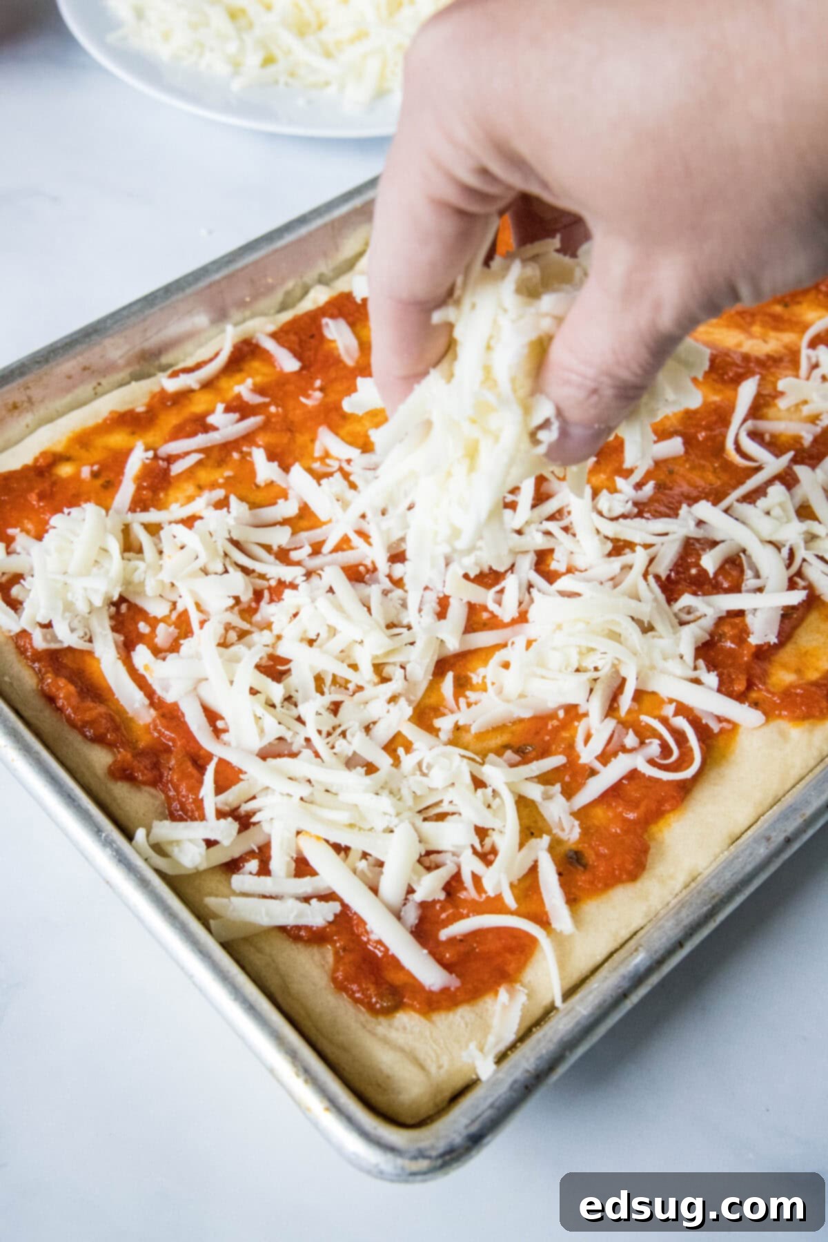 Cheesy crescent roll pizza is the perfect dinner or party snack! There’s zero kneading or rise time, just add your favorite toppings and bake! A hand sprinkling shredded mozzarella cheese over a crescent dough crust topped with pizza sauce.