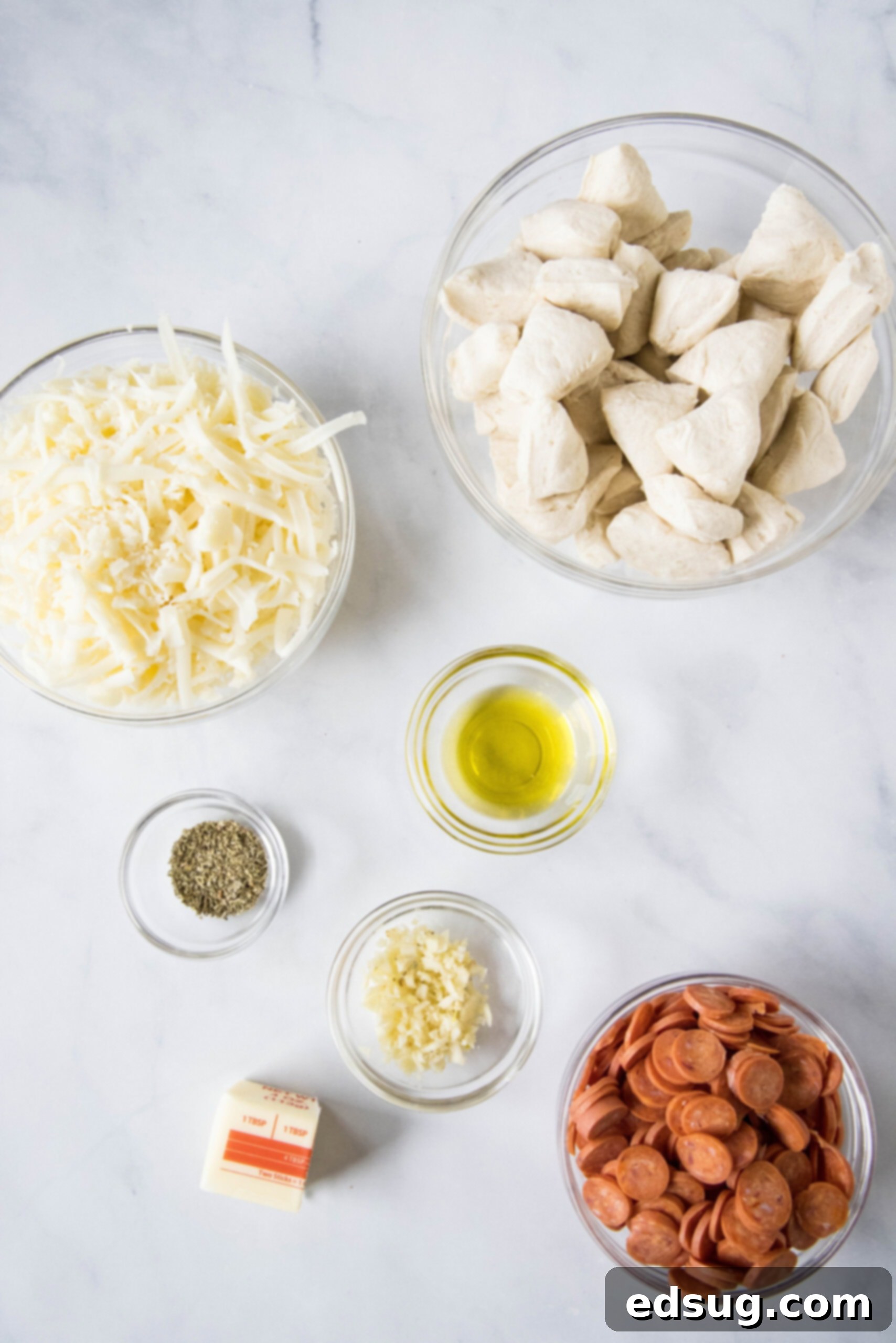 Pizza monkey bread is an easy, pull-apart-style appetizer or dinner made from puffy biscuits baked with pepperoni and lots of melty cheese! A collection of ingredients laid out for making pizza monkey bread, including biscuit dough, pepperoni, mozzarella, butter, olive oil, and seasonings.