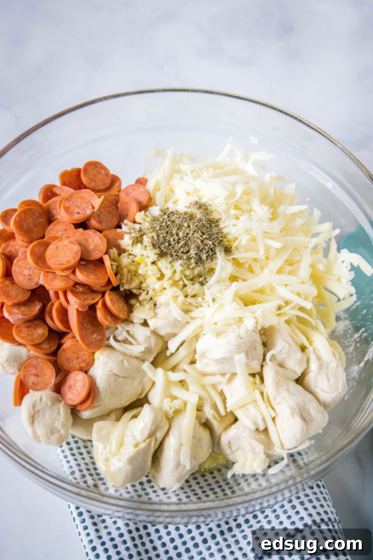 Pizza monkey bread is an easy, pull-apart-style appetizer or dinner made from puffy biscuits baked with pepperoni and lots of melty cheese! Biscuit dough pieces, pepperoni, mozzarella, and seasonings combined in a glass mixing bowl, coated and ready for baking.