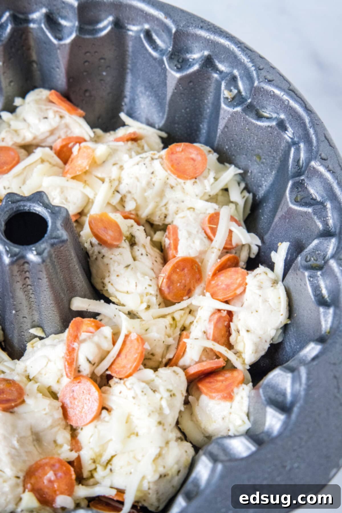 Pizza monkey bread is an easy, pull-apart-style appetizer or dinner made from puffy biscuits baked with pepperoni and lots of melty cheese! The combined biscuit dough balls and pizza ingredients neatly arranged in a greased bundt pan, ready for the oven.