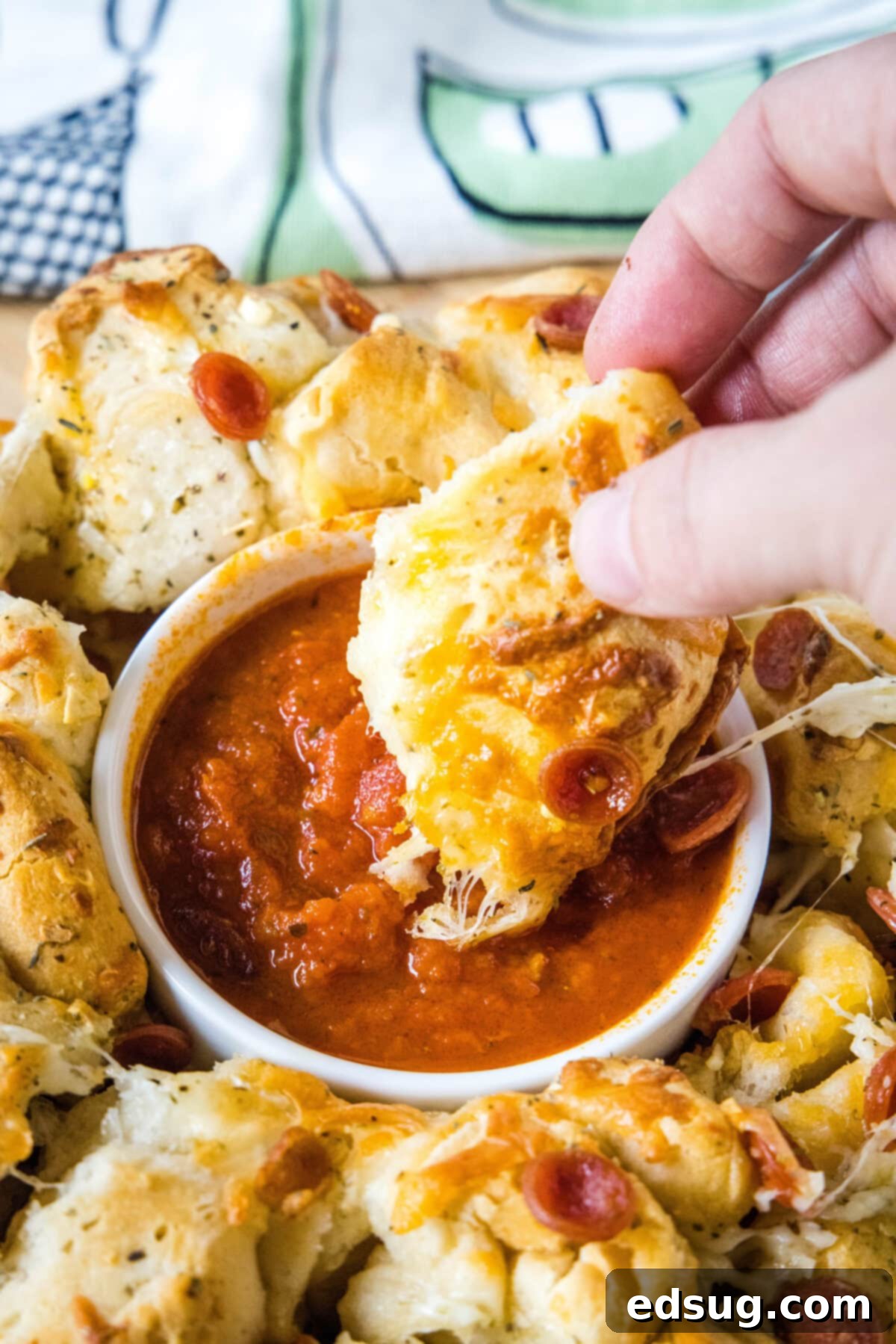 Pizza monkey bread is an easy, pull-apart-style appetizer or dinner made from puffy biscuits baked with pepperoni and lots of melty cheese! A hand pulling a piece of pizza monkey bread from the main loaf, ready for dipping into a bowl of marinara sauce.