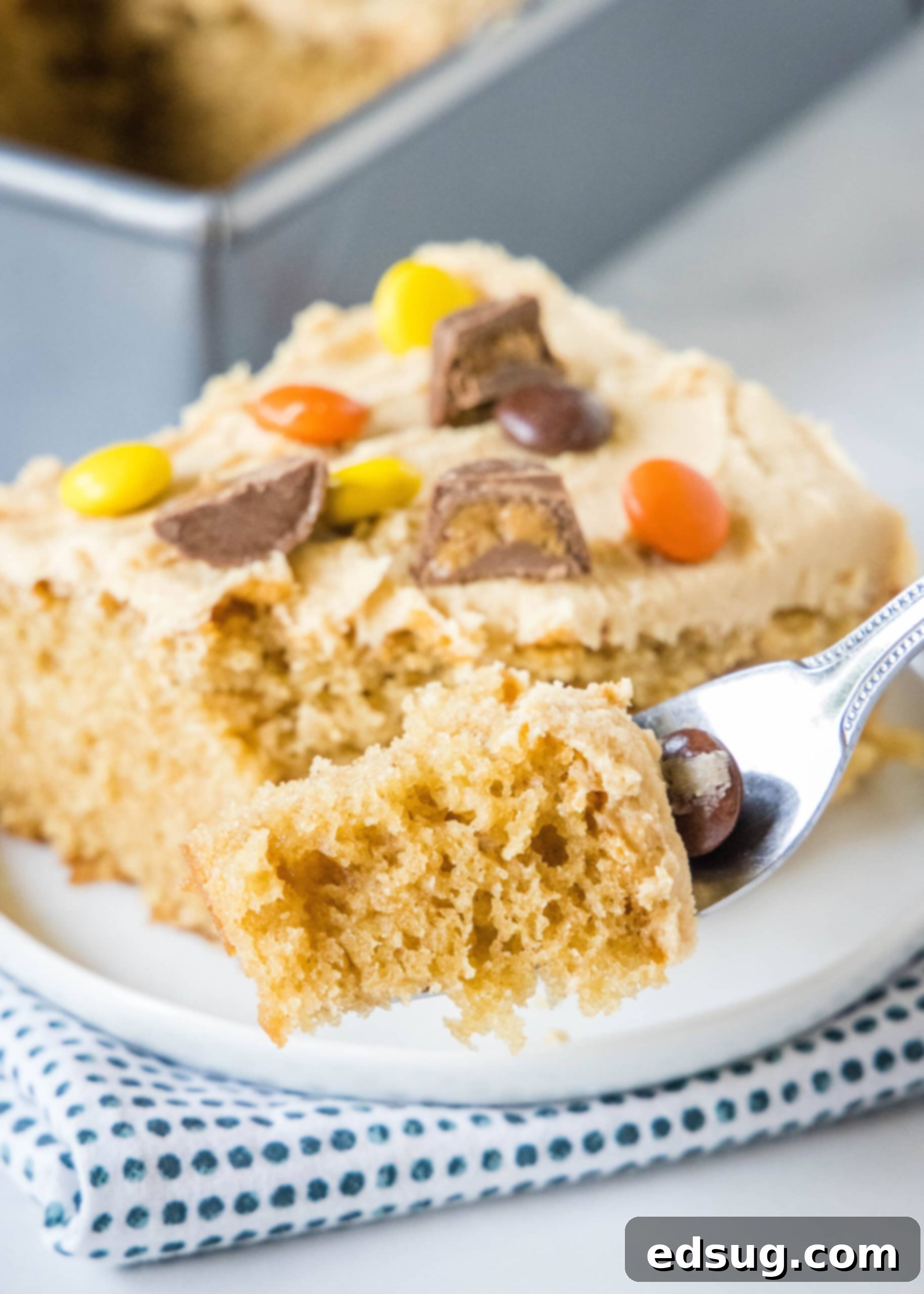 A fork lifts a bite of peanut butter cake with frosting, positioned in front of a full slice decorated with chopped peanut butter cups, highlighting its moist texture.