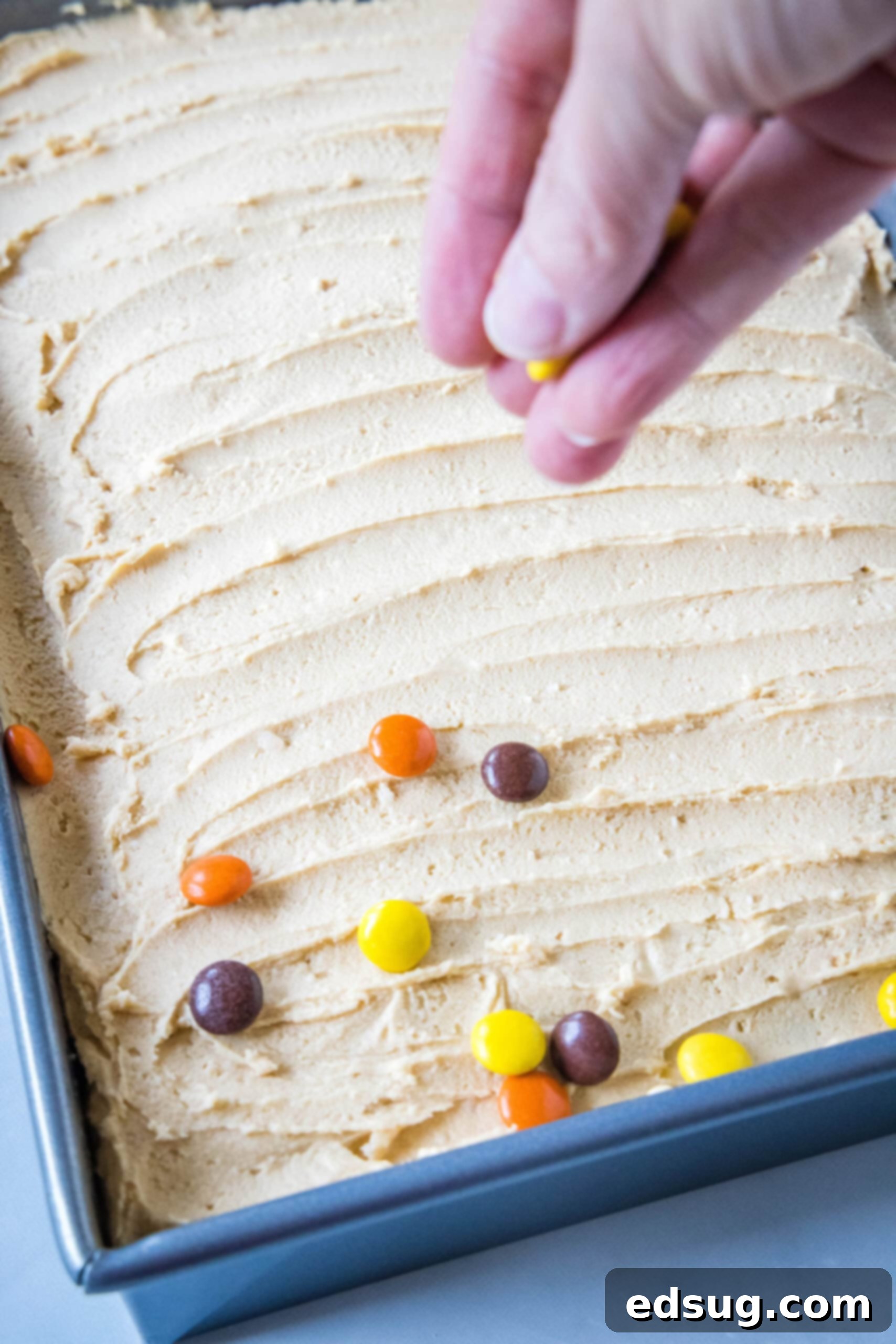 A hand gracefully sprinkling colorful Reese's pieces over a freshly frosted peanut butter cake, still in its sheet pan, adding a final decorative and flavorful touch.