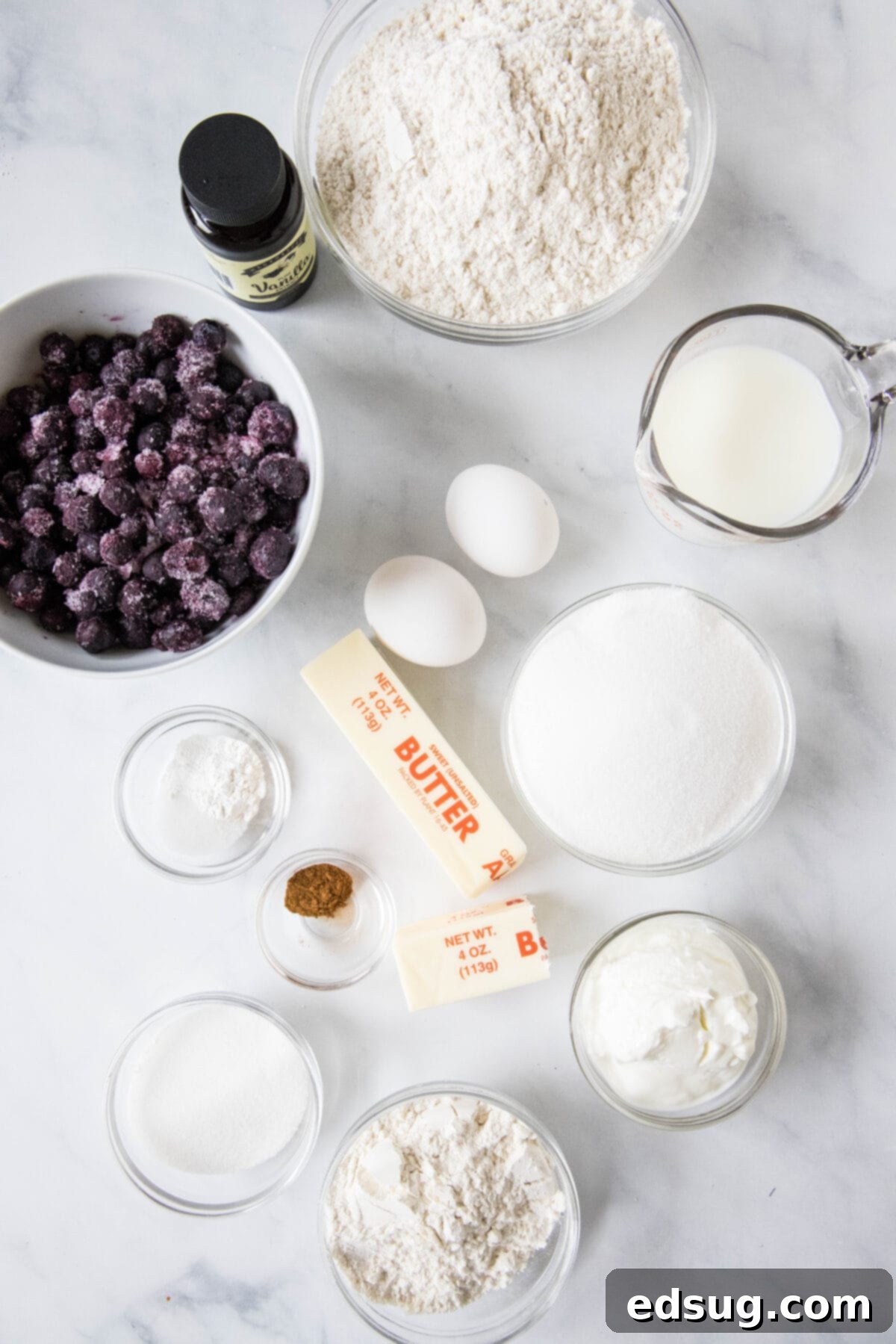Close-up of fresh, raw ingredients for making blueberry muffins: flour, butter, sugar, eggs, vanilla, Greek yogurt, and a bowl of frozen blueberries.