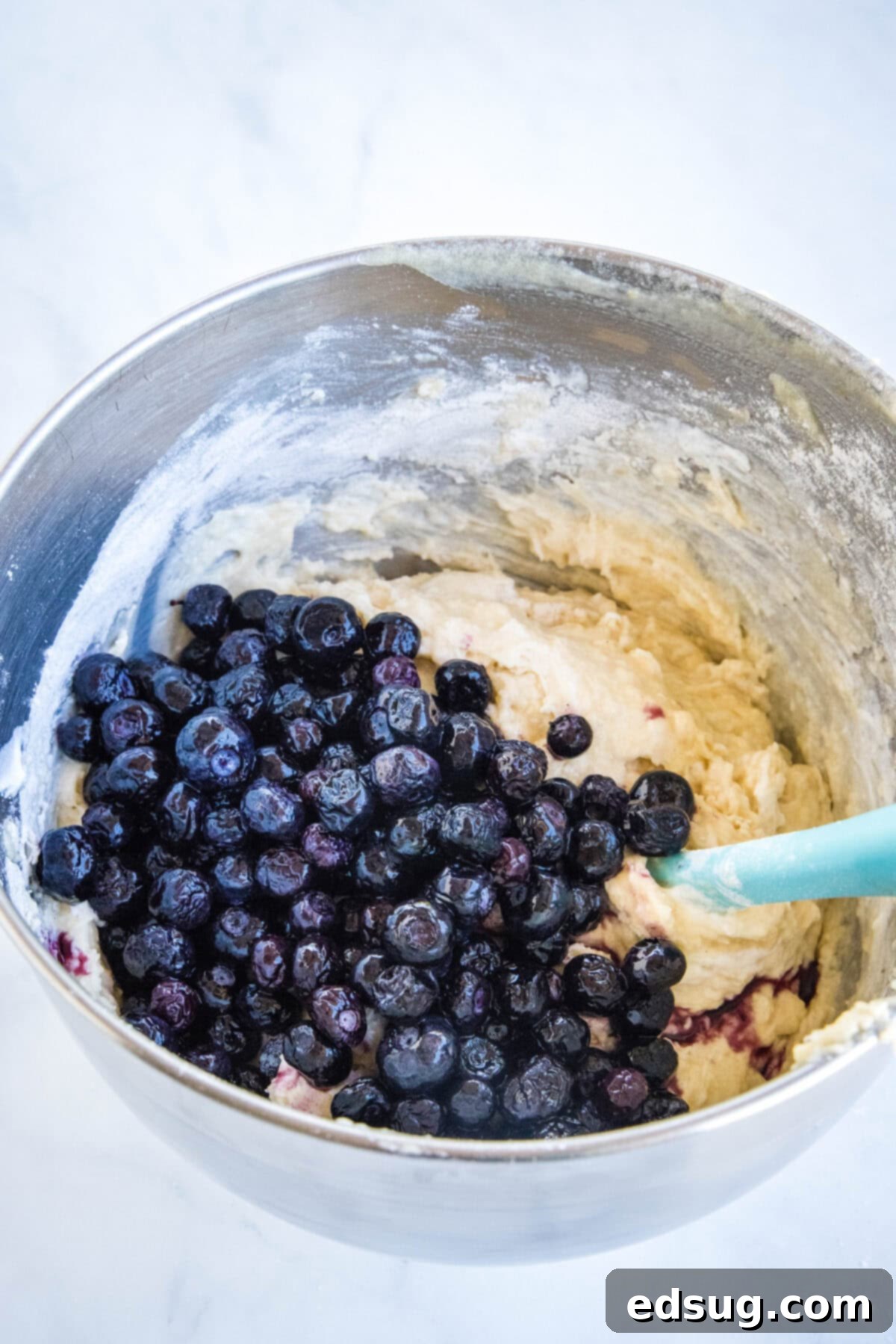 A bowl of thick, golden muffin batter with vibrant frozen blueberries gently folded into it, ready to be scooped.