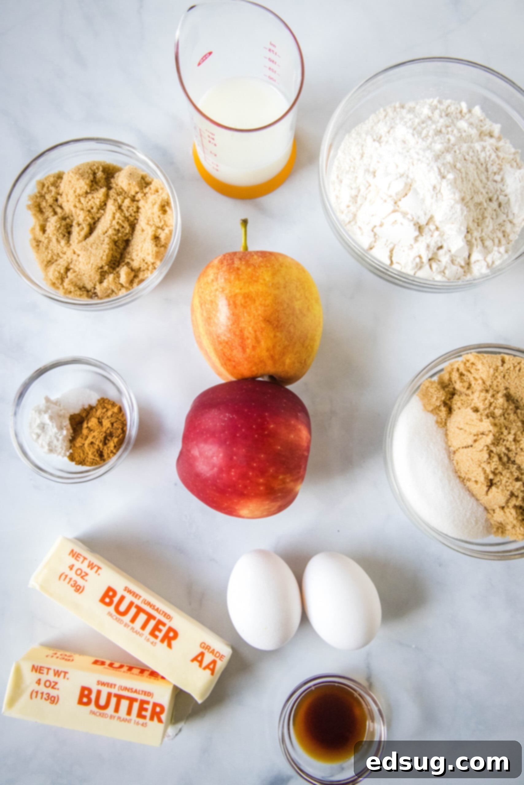 A vibrant selection of fresh ingredients laid out, featuring sliced apples, brown sugar, butter, cinnamon, vanilla extract, flour, baking powder, salt, granulated sugar, eggs, and milk, all ready for making a delicious apple upside down cake.