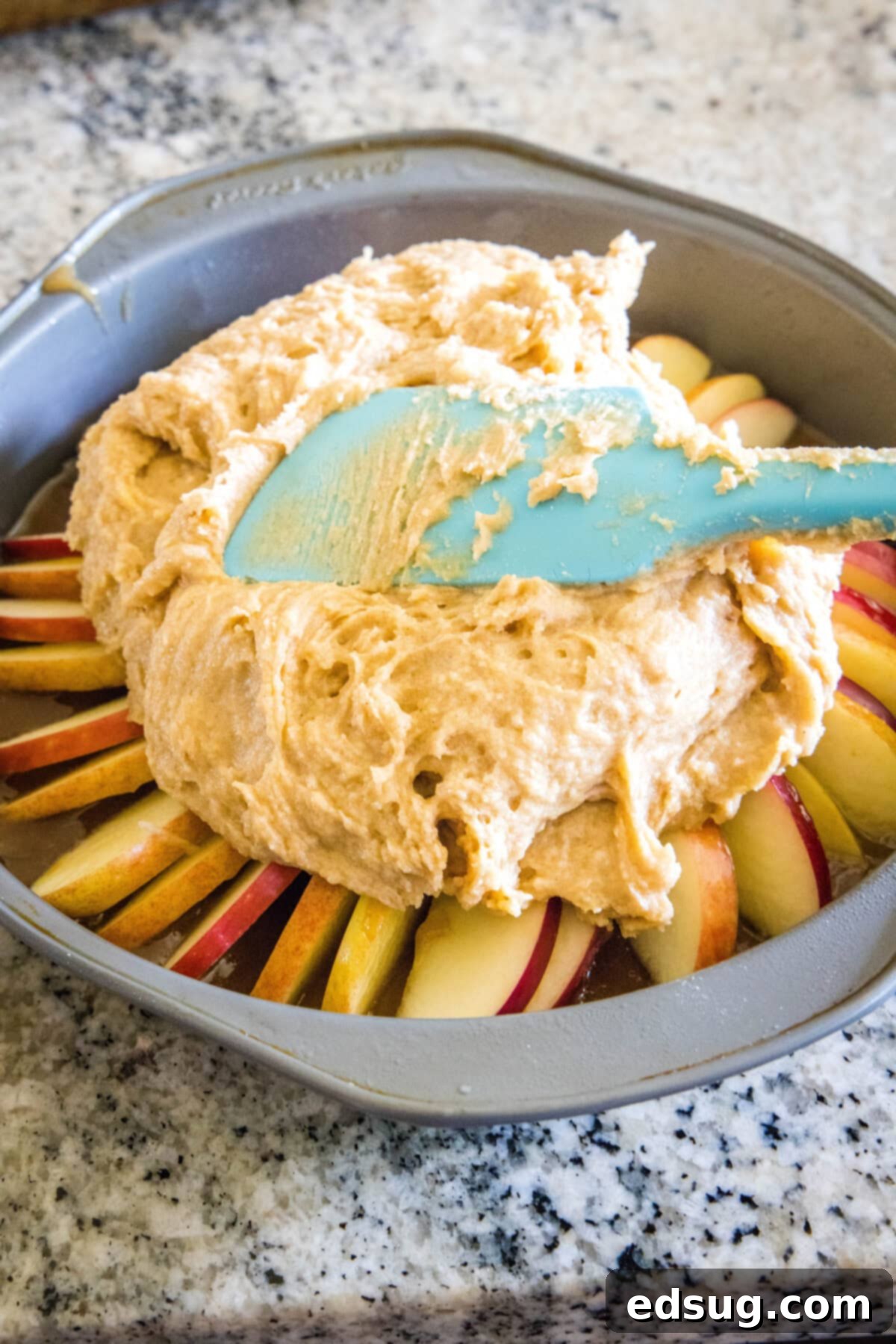 Smooth cake batter being carefully spread in an even layer over the organized caramel apple base within a round cake pan, ready for baking.