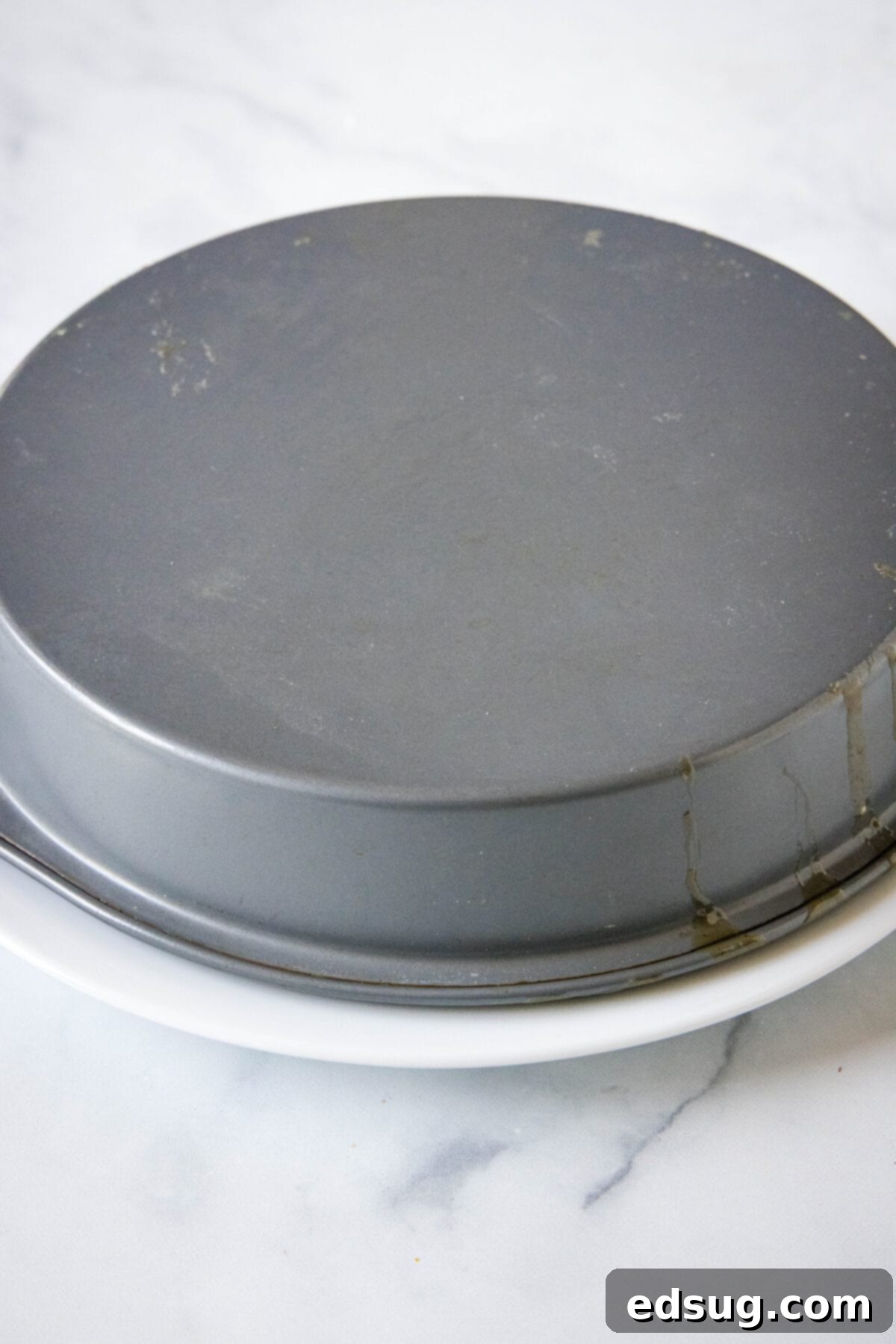 A round baking pan inverted upside down onto a white serving plate, depicting the crucial moment of flipping the cake to reveal its caramelized apple topping.
