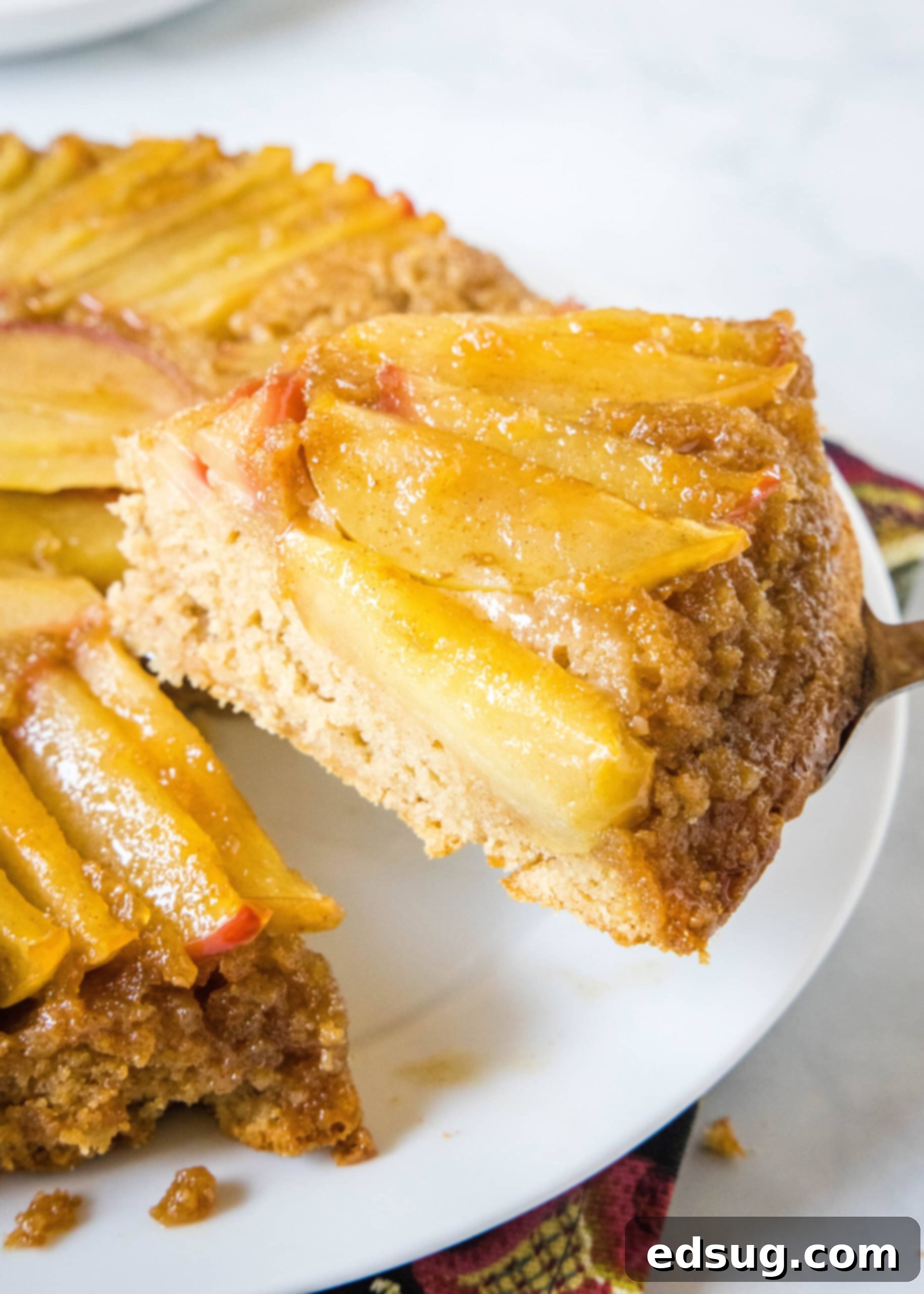 A close-up view of a delicious slice of apple upside down cake being carefully lifted away from the rest of the cake on a white plate, showcasing its moist texture and caramelized apple topping.