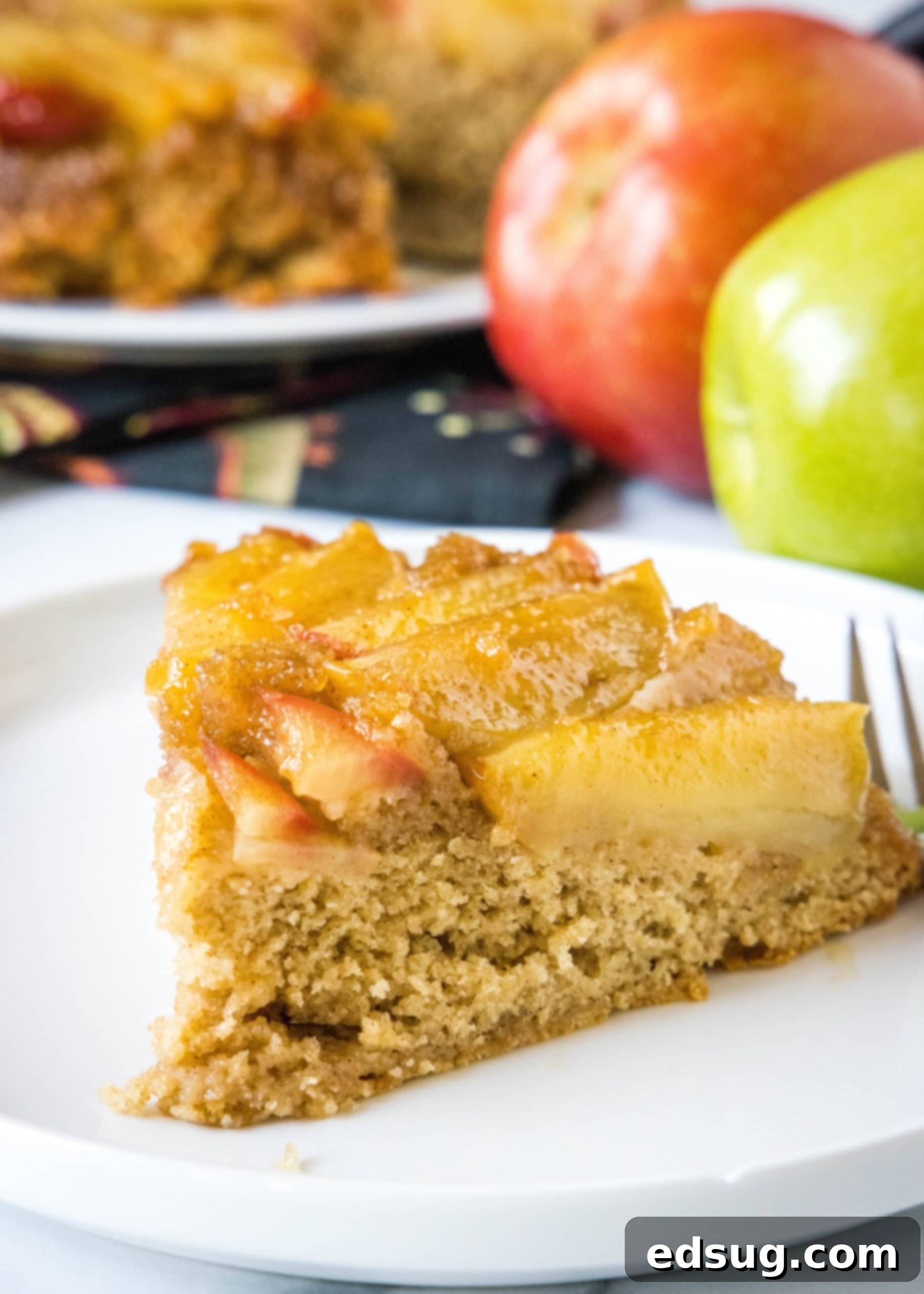 A charming slice of apple upside down cake presented on a white plate next to a fork, with fresh red and green apples artfully blurred in the background, hinting at the cake's main ingredient.