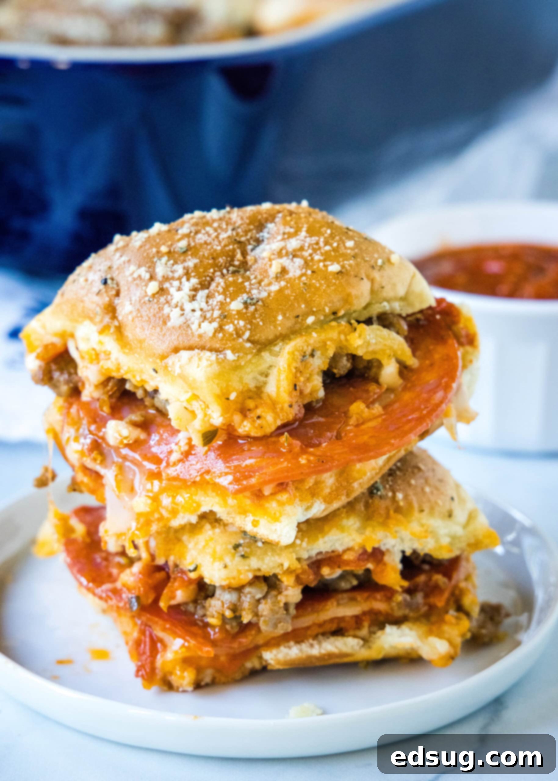 Hawaiian roll pizza sliders are a fun appetizer or weeknight family dinner! They're quick and easy to make with your favorite pizza toppings. Two golden-brown Hawaiian roll pizza sliders stacked on a white plate, with melted cheese, pepperoni visible, and a small bowl of marinara sauce in the soft-focus background, ready for dipping. The rolls are fluffy and the toppings are perfectly integrated.