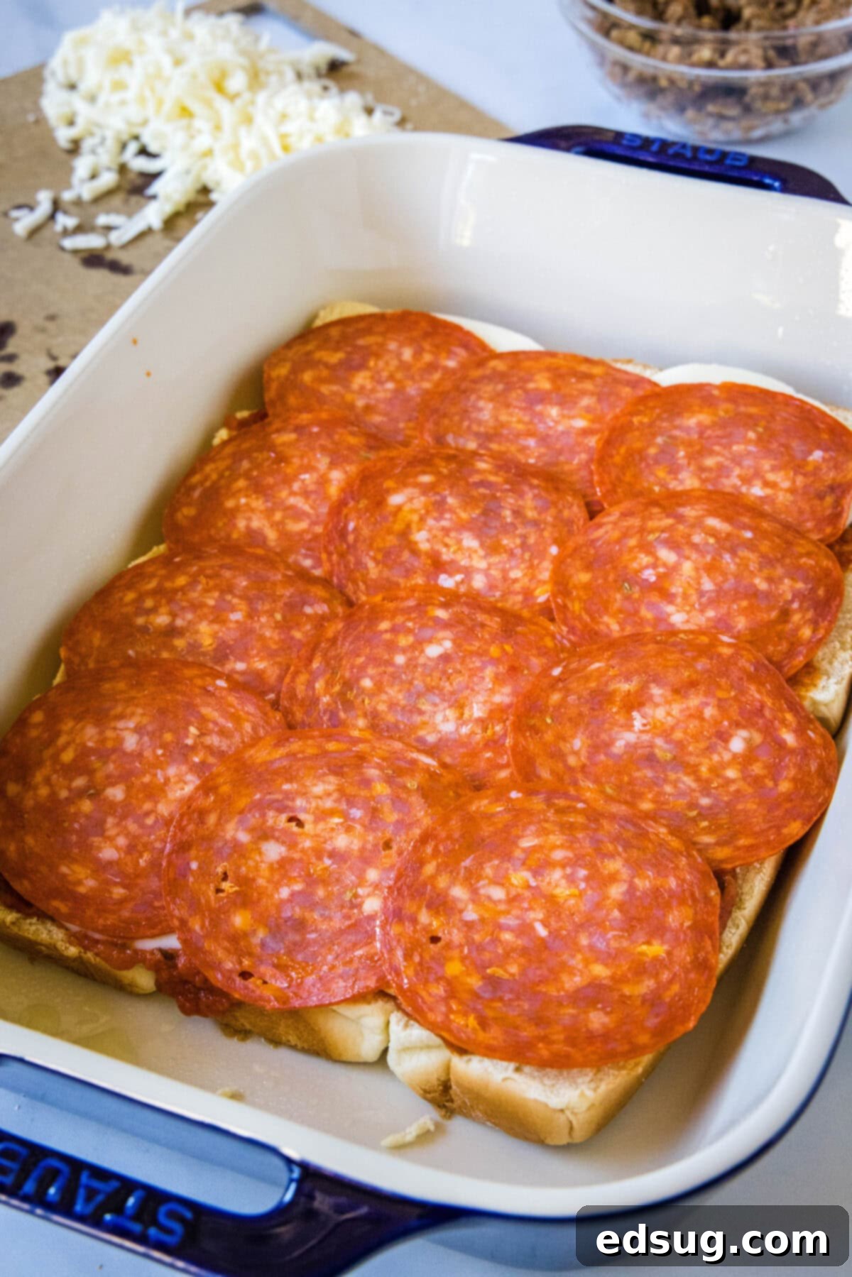 Hawaiian roll pizza sliders are a fun appetizer or weeknight family dinner! They're quick and easy to make with your favorite pizza toppings. Slices of bright red pepperoni being carefully arranged and layered over the bottom halves of soft, sweet Hawaiian rolls in a greased ceramic baking dish. The rolls are still connected, forming a cohesive base for the pizza sliders.