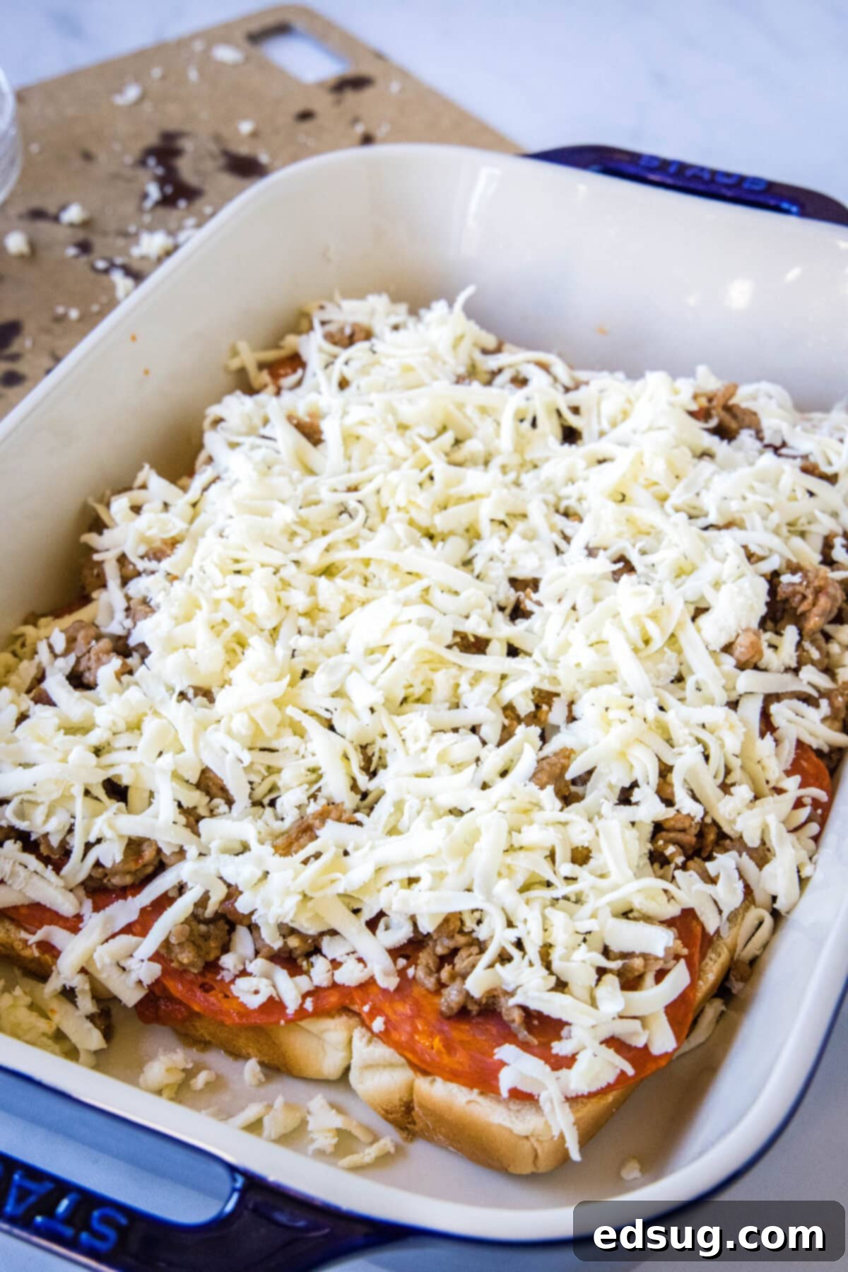 Hawaiian roll pizza sliders are a fun appetizer or weeknight family dinner! They're quick and easy to make with your favorite pizza toppings. A comprehensive view of the assembled bottom half of the pizza sliders in a blue baking dish, showcasing the layers of rich pizza sauce, sliced provolone cheese, savory pepperoni, crumbled Italian sausage, and a generous topping of shredded cheddar cheese, all before the top buns are added.