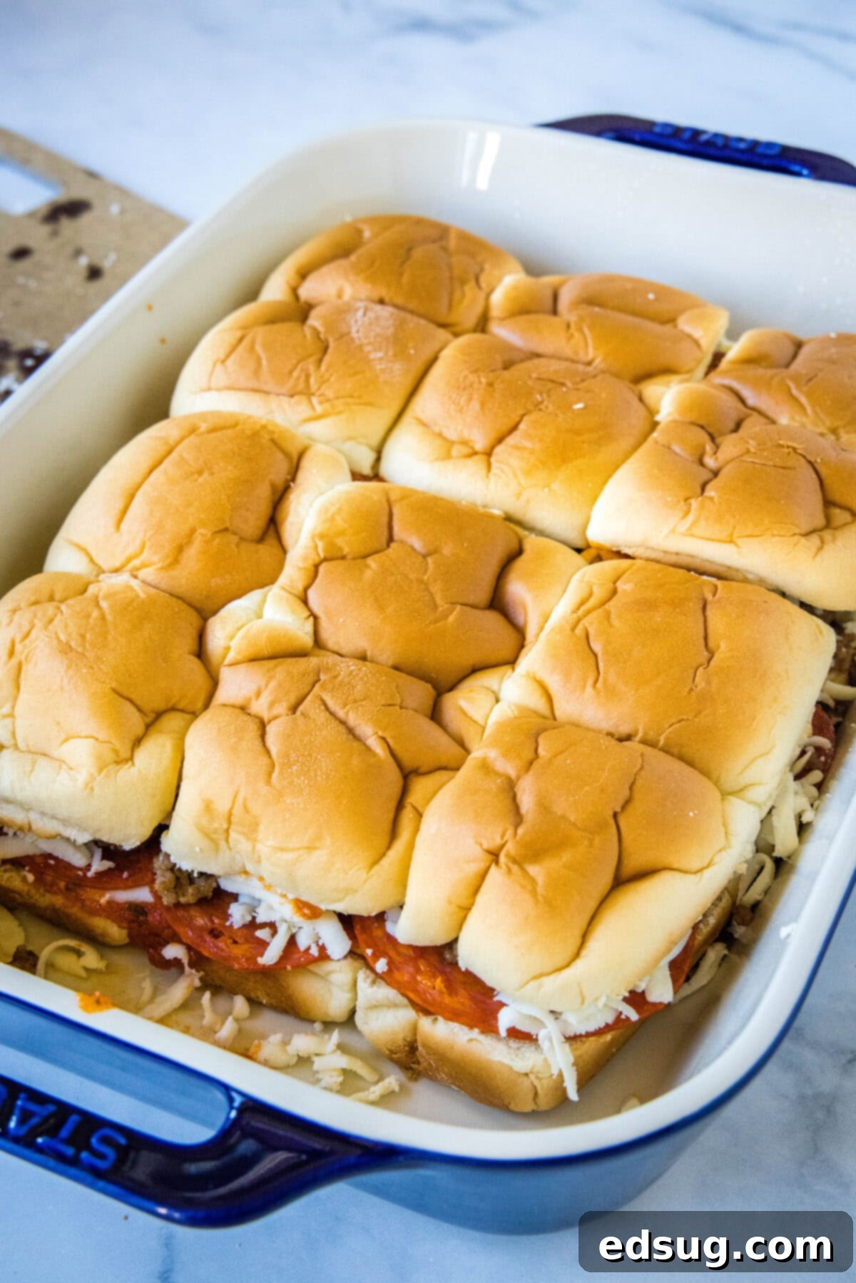 Hawaiian roll pizza sliders are a fun appetizer or weeknight family dinner! They're quick and easy to make with your favorite pizza toppings. The fully assembled Hawaiian roll pizza sliders, with all layers including the top buns and preliminary garlic butter, nestled neatly in a blue ceramic baking dish, prior to being placed in the oven for baking. The composition is clean and inviting.