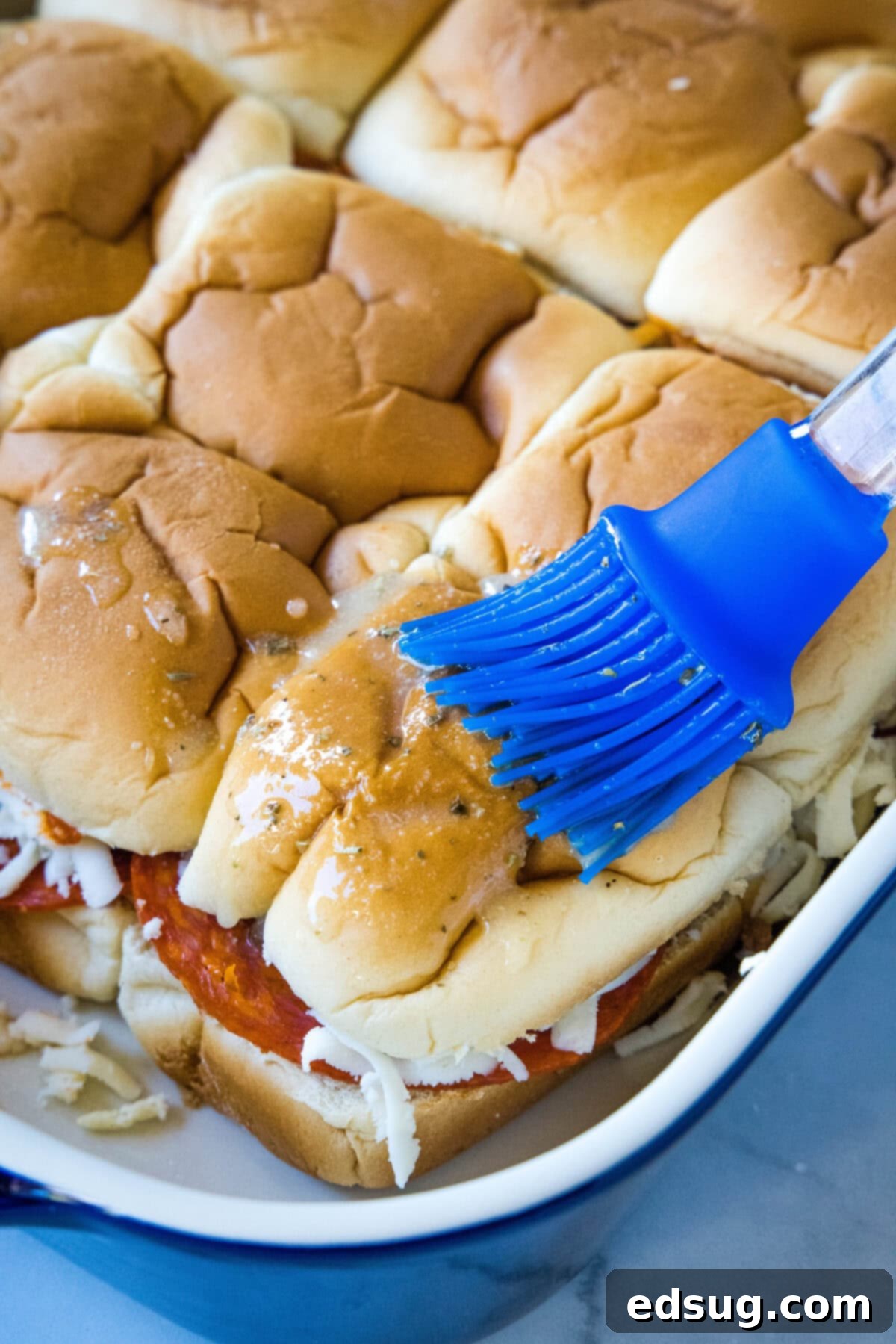 Hawaiian roll pizza sliders are a fun appetizer or weeknight family dinner! They're quick and easy to make with your favorite pizza toppings. A blue basting brush delicately applies the savory garlic butter mixture over the tops of the assembled pizza sliders in a baking dish. The melted butter glistens on the fluffy Hawaiian rolls, enhancing their potential for a golden, flavorful crust during baking.