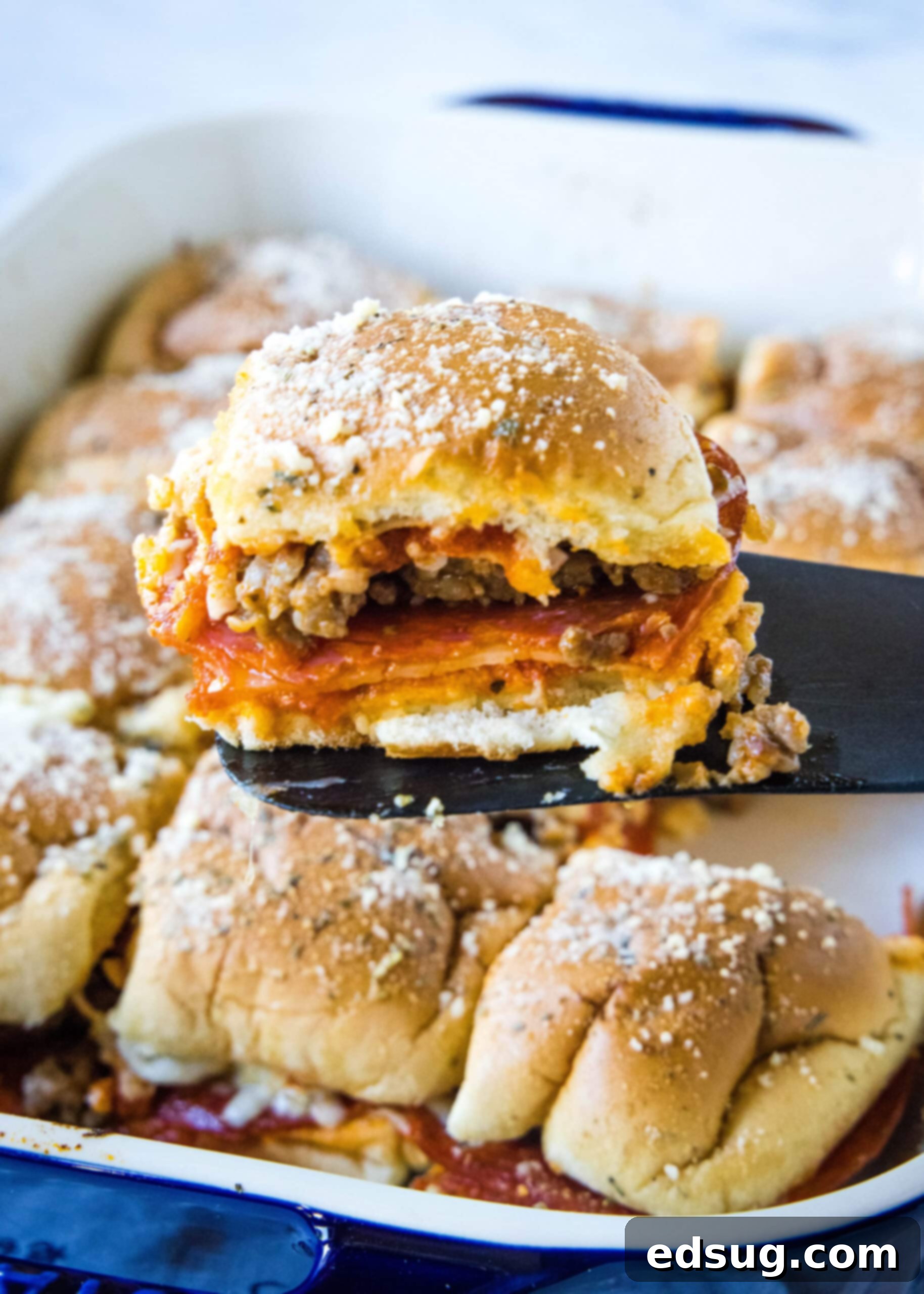Hawaiian roll pizza sliders are a fun appetizer or weeknight family dinner! They're quick and easy to make with your favorite pizza toppings. A single, perfectly golden-brown and cheesy pizza slider is being carefully lifted from a blue ceramic baking dish using a black spatula. Melted cheese stretches slightly, and the golden crust of the Hawaiian roll is visible, indicating it's ready to be served and enjoyed.