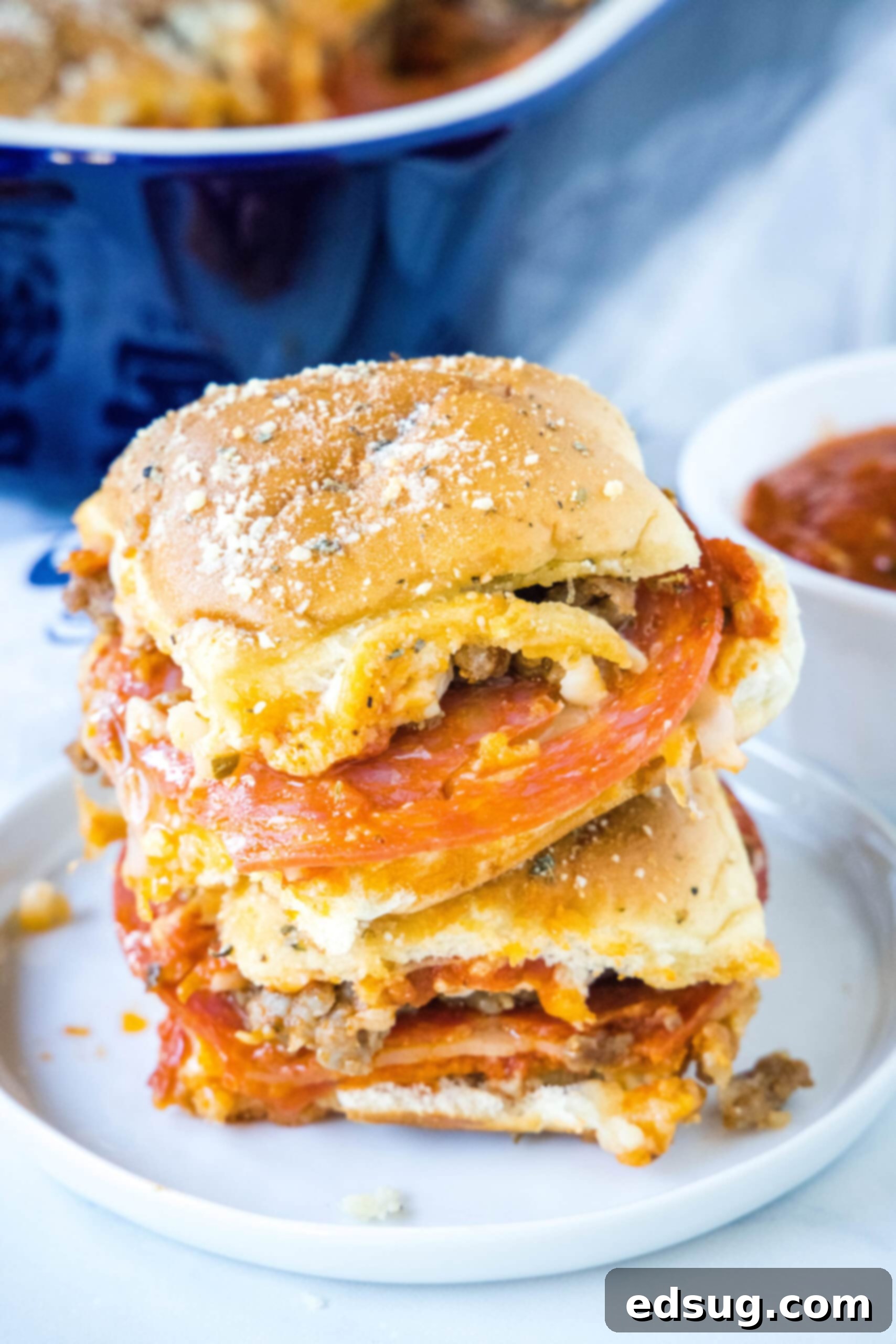 Hawaiian roll pizza sliders are a fun appetizer or weeknight family dinner! They're quick and easy to make with your favorite pizza toppings. Two fully baked, golden-brown pizza sliders with melted cheese and pepperoni are stacked neatly on a white ceramic plate, positioned slightly to the left. In the soft-focus background, a small white bowl filled with vibrant red marinara sauce and a sprig of green herbs waits, suggesting a perfect dipping accompaniment.