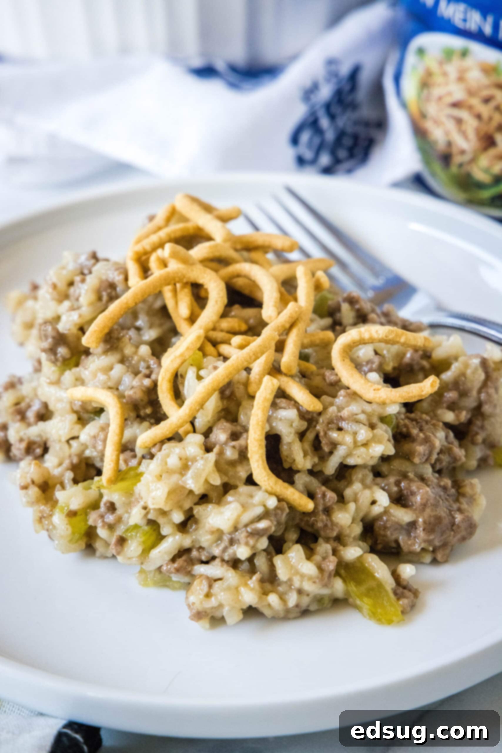 Hamburger rice casserole is an easy retro dinner recipe with fluffy rice, juicy ground beef, and veggies cooked in cream of mushroom soup! A serving of hamburger rice casserole topped with dried chow mein noodles next to a fork on a white plate.