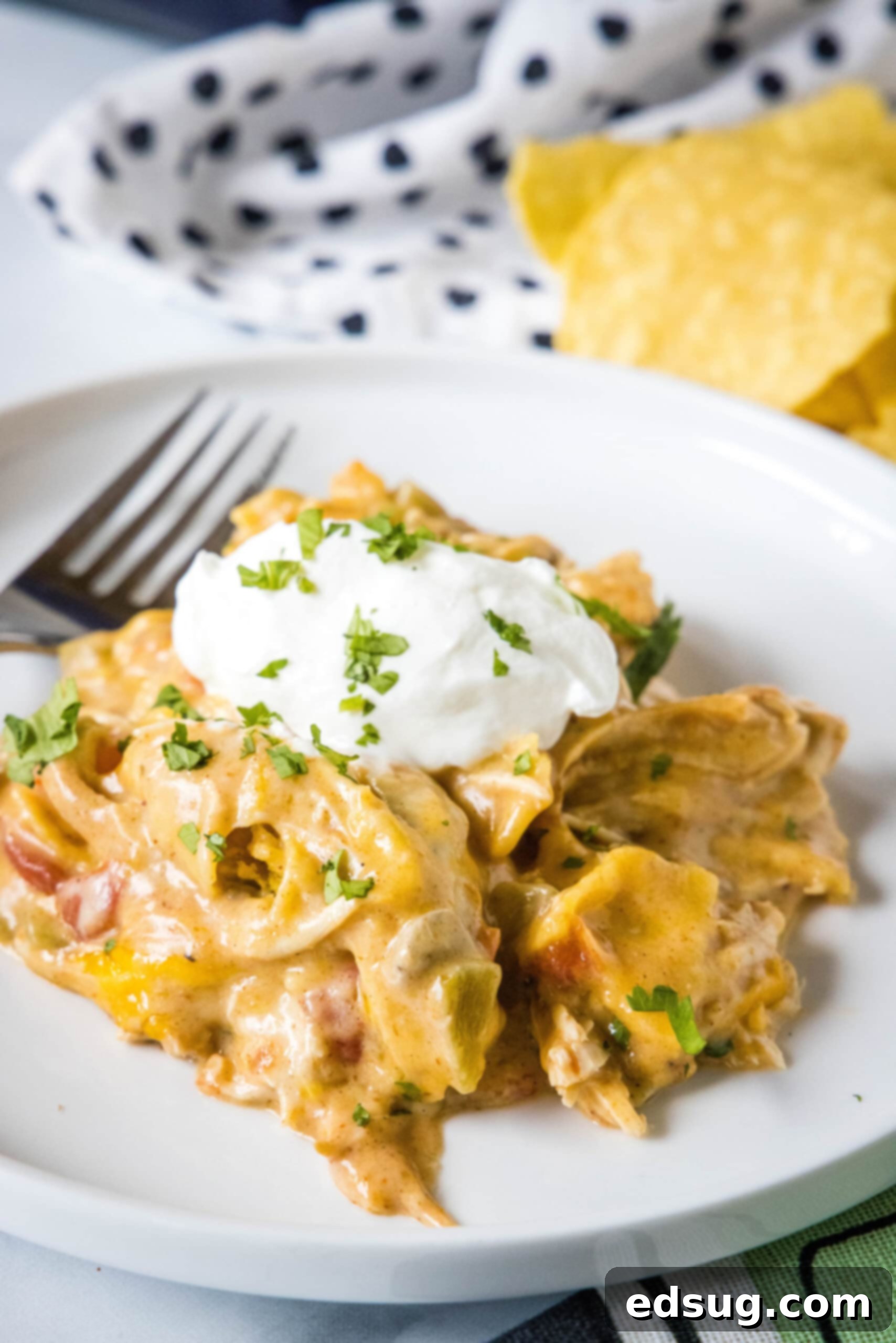 A serving of King Ranch chicken casserole topped with a dollop of sour cream on a white plate next to a fork, showcasing its cheesy, layered goodness.