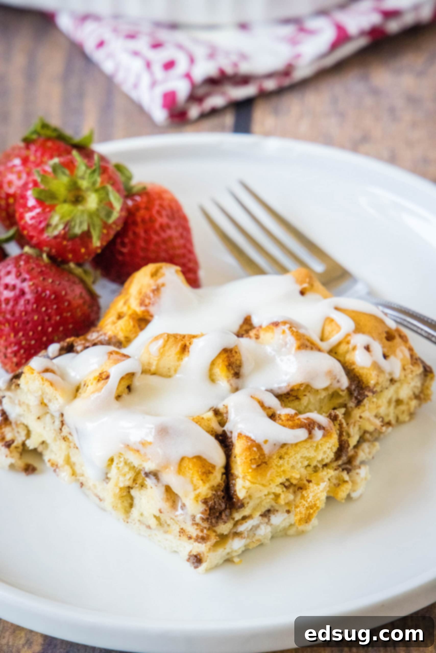 Cinnamon roll casserole is a quick and easy breakfast casserole with flaky layers baked in a rich and creamy custard. Perfect for a crowd! A serving of cinnamon roll casserole topped with glaze, next to a fork and fresh strawberries on a white plate, highlighting its deliciousness and simple elegance.