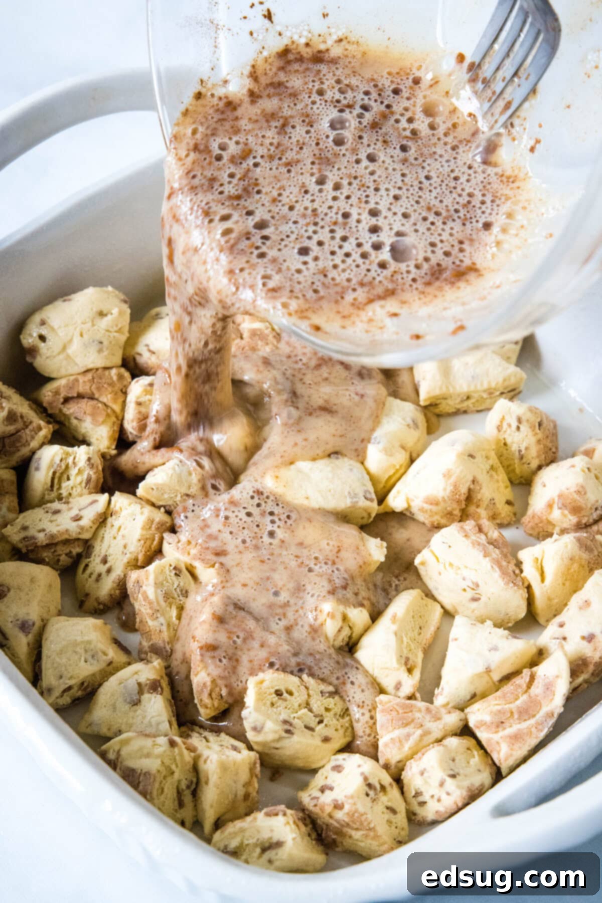 Cinnamon roll casserole is a quick and easy breakfast casserole with flaky layers baked in a rich and creamy custard. Perfect for a crowd! Creamy vanilla custard being poured evenly over cut cinnamon roll pieces arranged in a ceramic baking dish, preparing for baking.