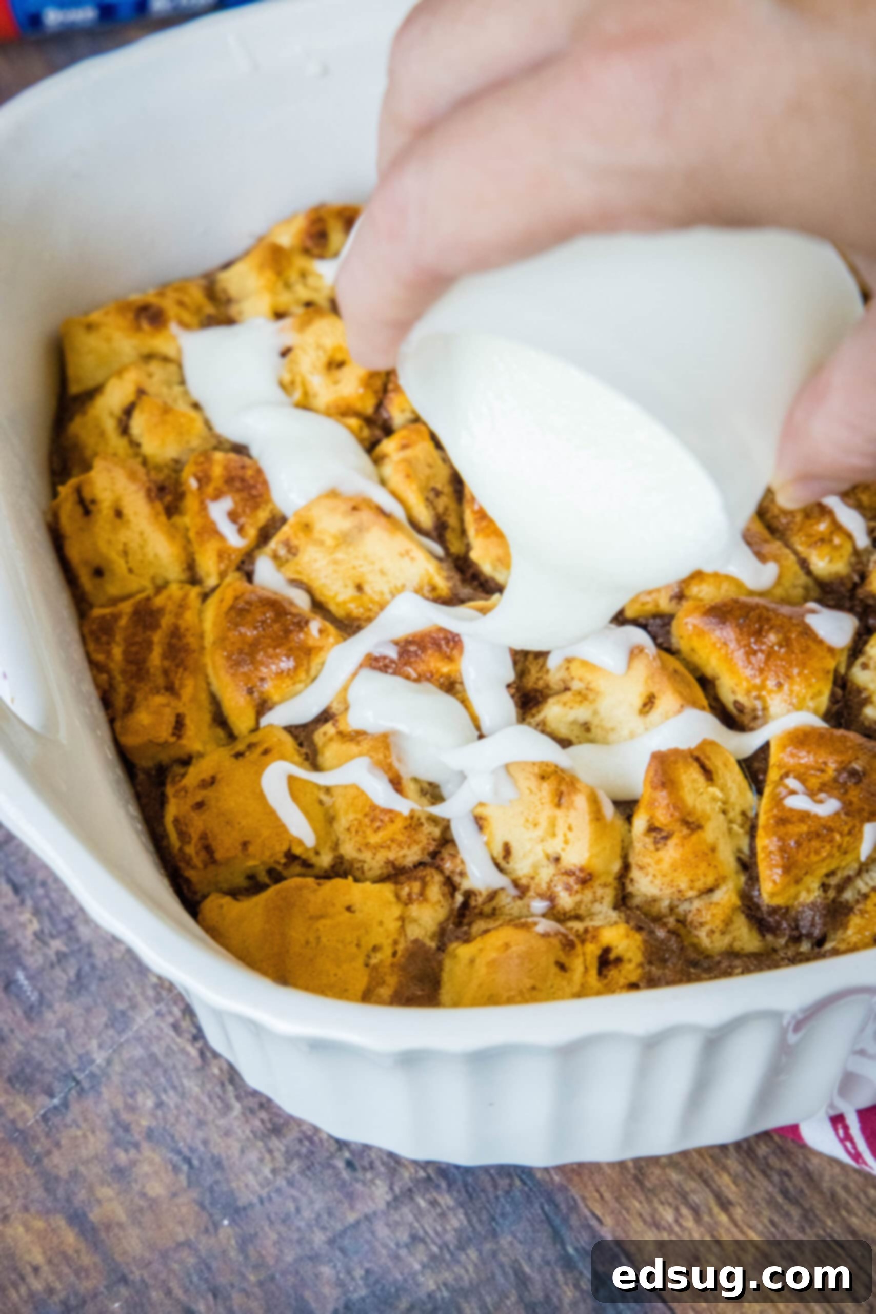 Cinnamon roll casserole is a quick and easy breakfast casserole with flaky layers baked in a rich and creamy custard. Perfect for a crowd! A hand carefully drizzling the sweet white icing over a warm, golden-baked cinnamon roll casserole in a ceramic dish, ready for serving.
