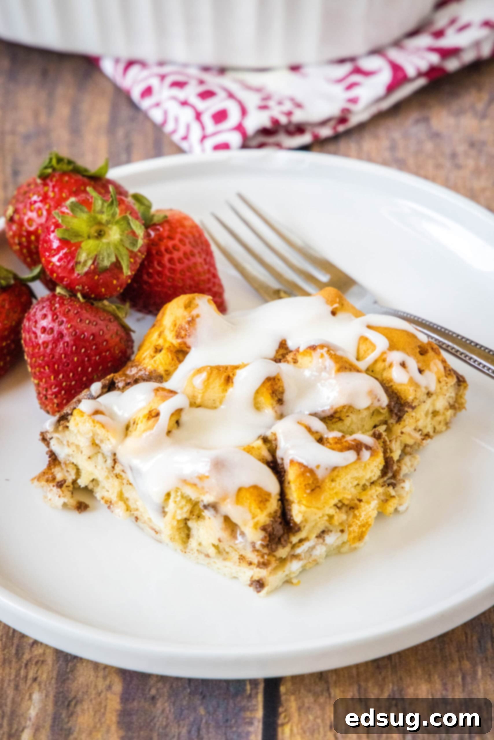 Cinnamon roll casserole is a quick and easy breakfast casserole with flaky layers baked in a rich and creamy custard. Perfect for a crowd! A perfectly portioned serving of cinnamon roll casserole, glistening with glaze, presented on a white plate with a fork and fresh strawberries, inviting indulgence.