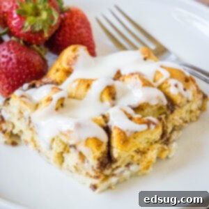 Ultimate Cinnamon Bliss Bake 2 A serving of cinnamon roll casserole topped with glaze, next to a fork and fresh strawberries on a white plate.