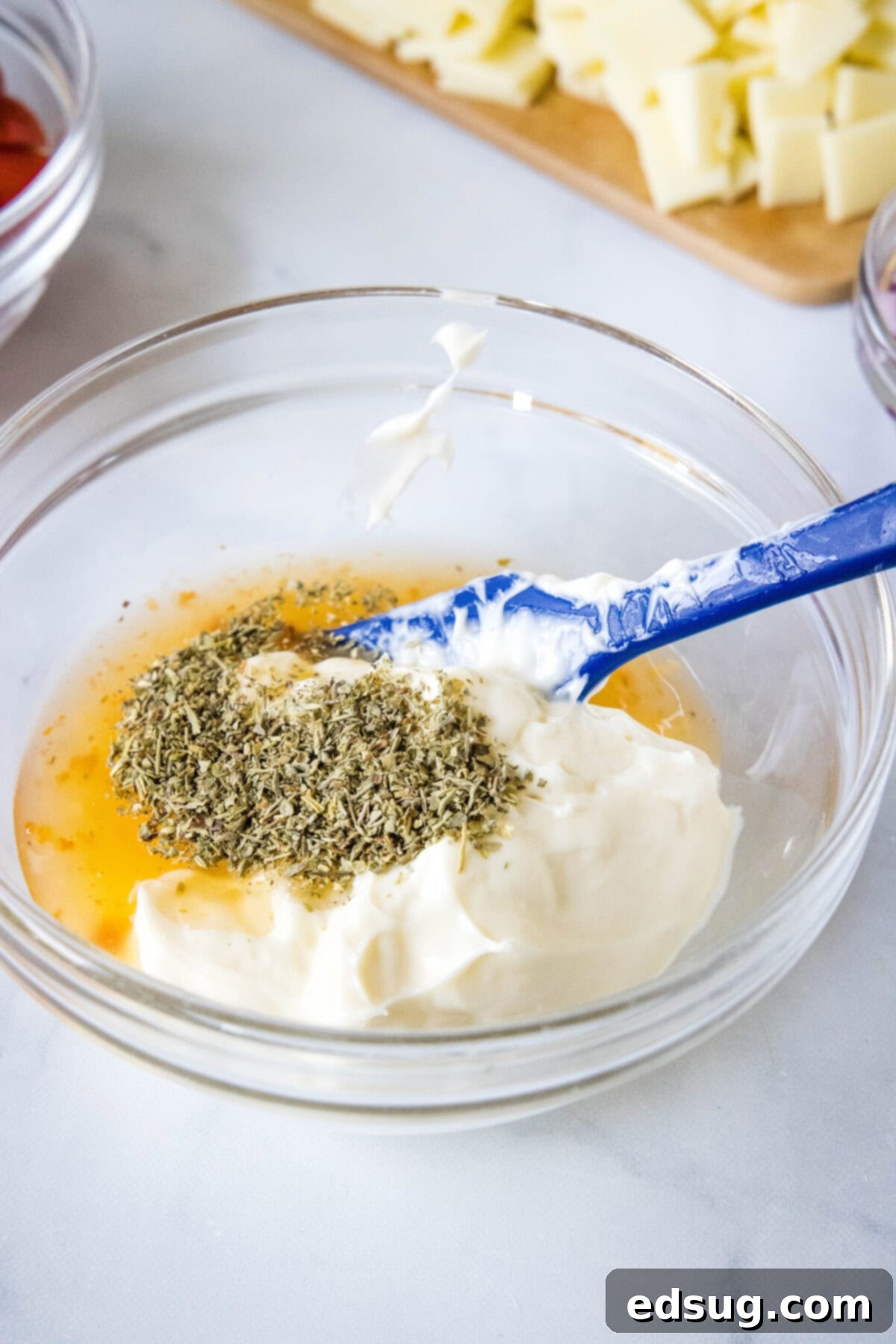 This fresh and zesty hoagie dip has all the flavors of an Italian sub sandwich! Serve this dip as an easy appetizer spread over crusty bread. Dressing ingredients added to a glass bowl with a blue spatula for stirring.