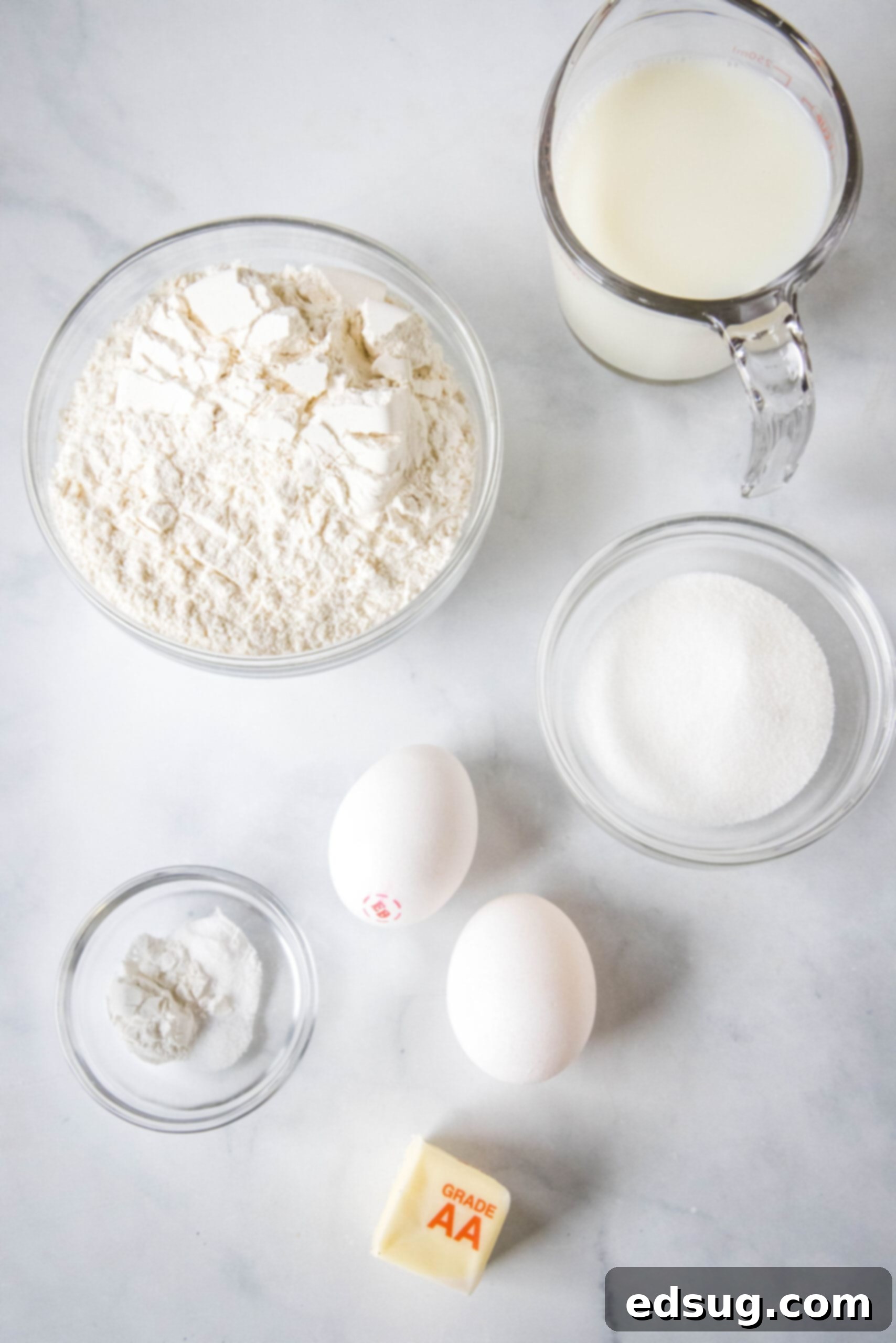 A collection of fresh ingredients for griddle cakes laid out on a kitchen counter, including flour, sugar, eggs, milk, and butter.