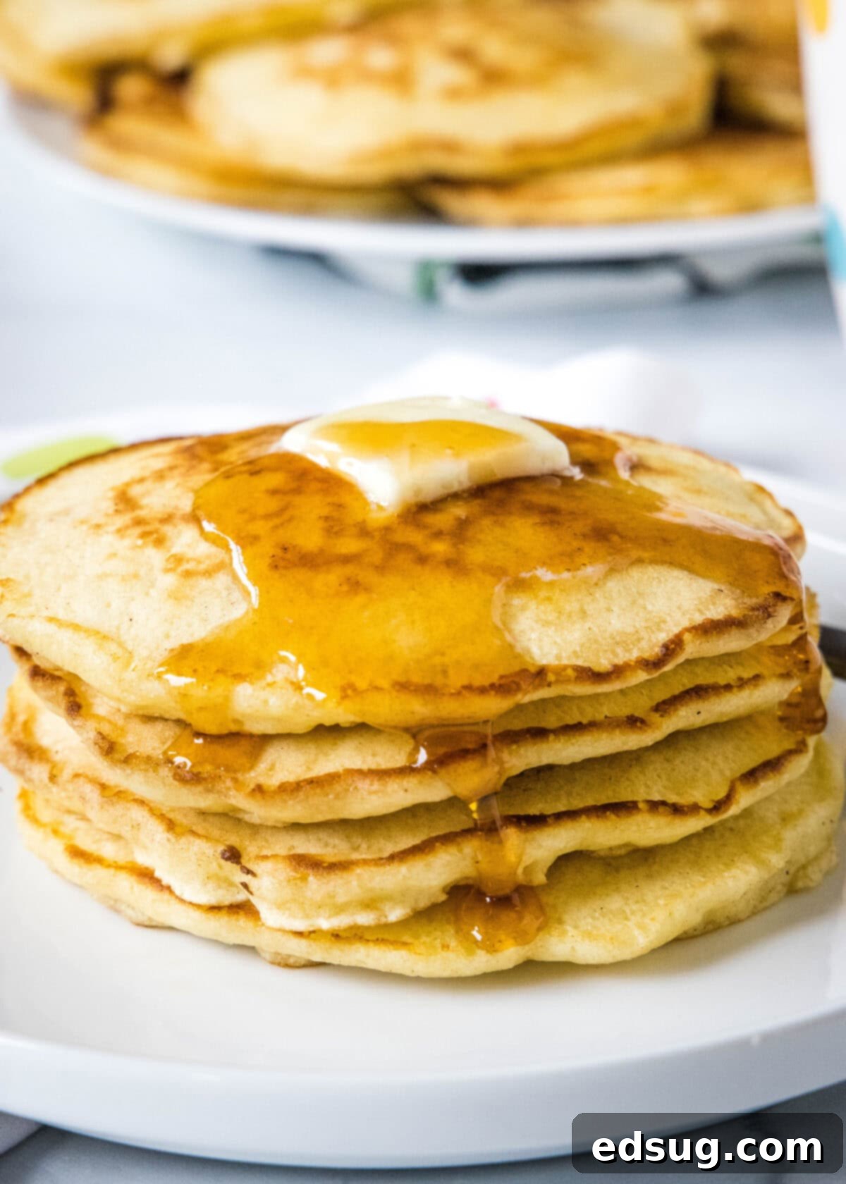 A stacked trio of griddle cakes, crowned with a melting pat of butter and drizzled with maple syrup, with more griddle cakes blurred in the background.