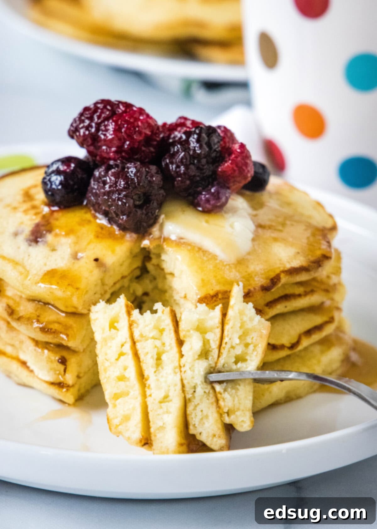 A fork piercing a piece of griddle cake, held in front of a stack adorned with mixed berries and maple syrup on a white plate, inviting a bite.