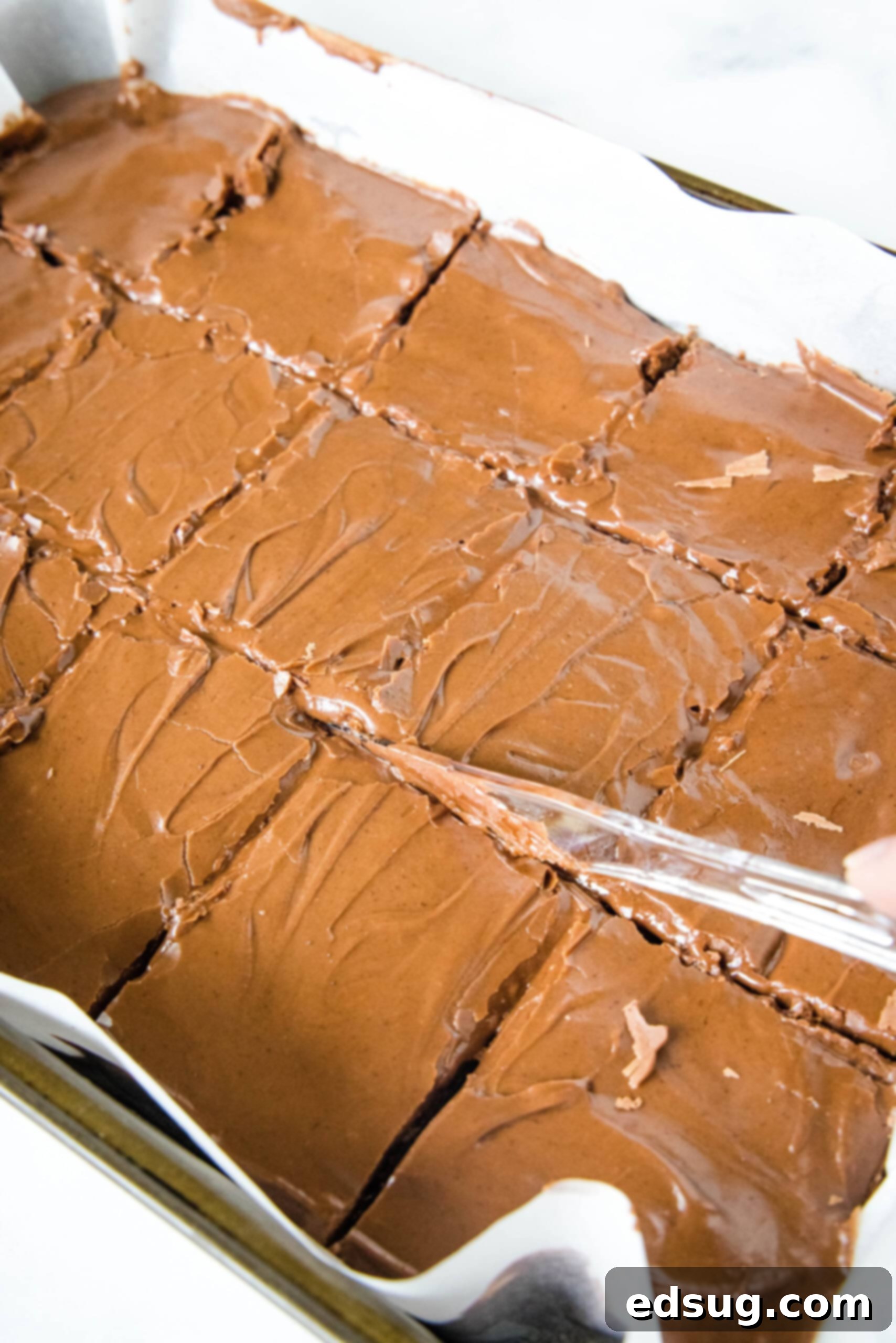 Lunch lady brownies are moist, fudgy brownies just like the ones served at school lunches! Every bite is filled with chocolate flavor. A hand using a plastic knife to cut a pan full of chocolate lunch lady brownies into neat squares, showing the fudgy texture and glossy frosting.
