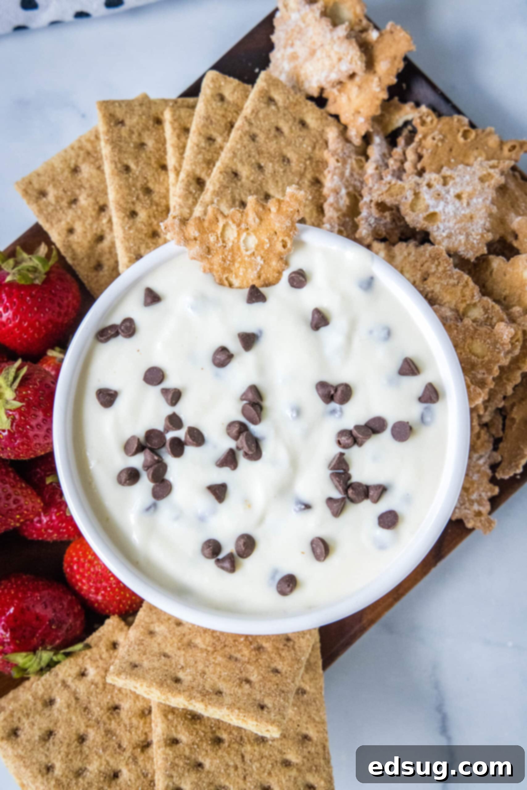 An overhead view of a beautifully presented cannoli dip bowl, garnished with chocolate chips and a graham cracker, on a rustic wooden platter.