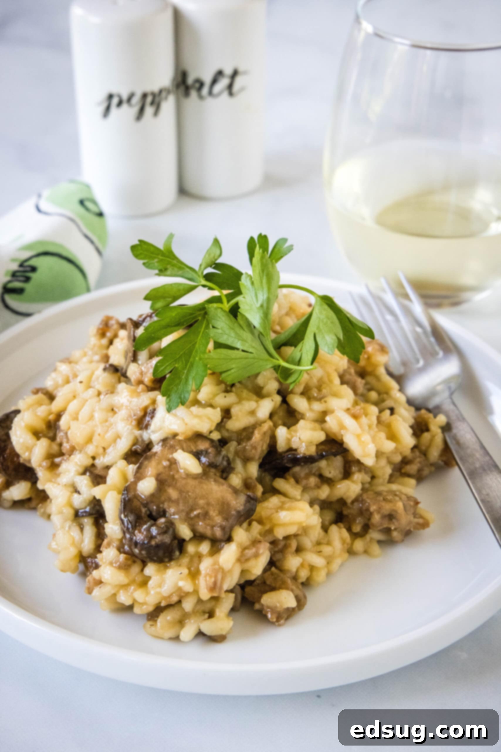 Sausage risotto garnished with fresh parsley on a white plate next to a fork, with a glass of wine next to salt and pepper shakers in the background.