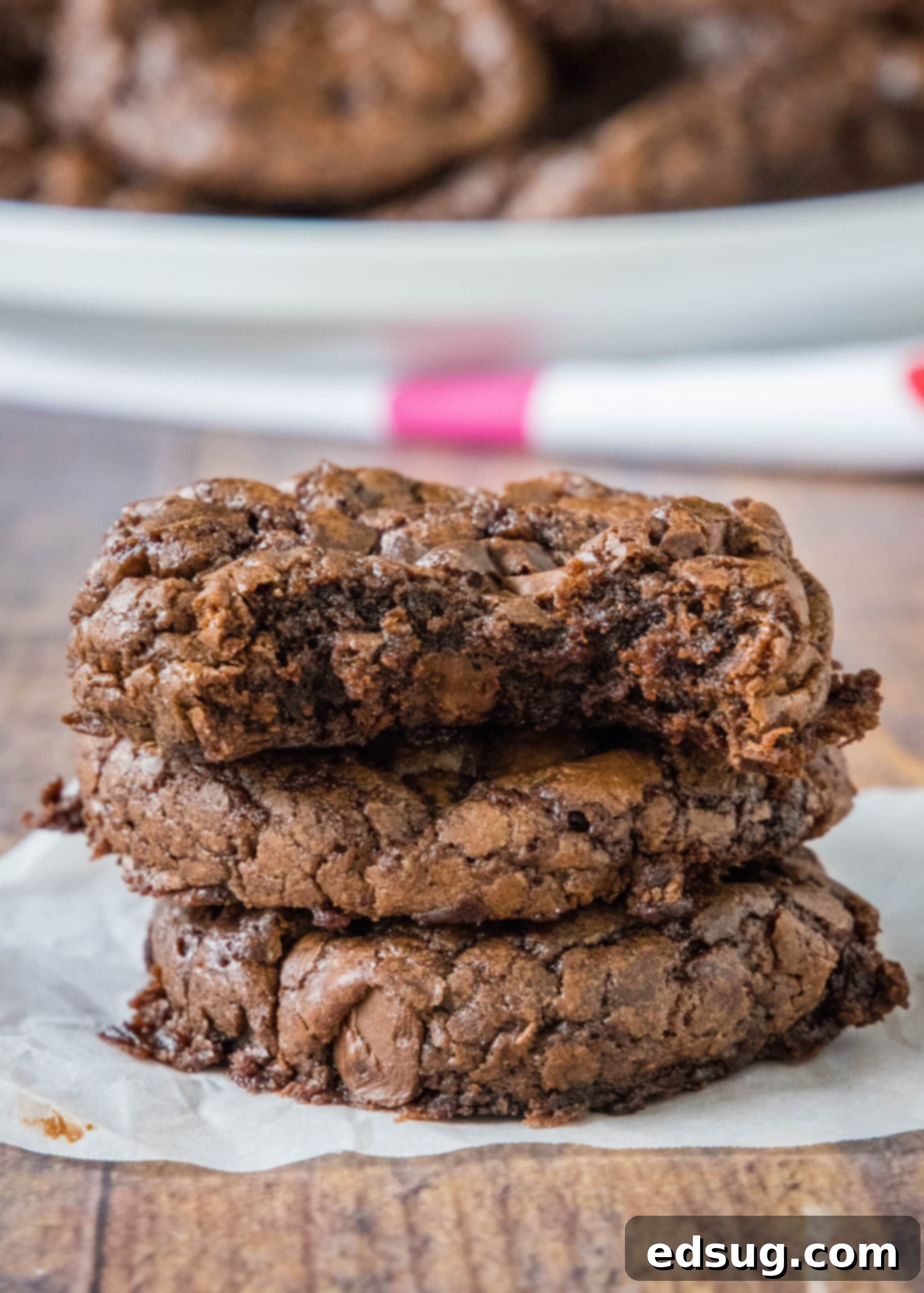 Brownie mix cookies are soft-baked, fudgy cookies made with only 4 ingredients! All you need is a box of brownie mix, oil, eggs, and flour. A stack of three rich, fudgy brownie mix cookies on parchment paper, with a bite taken out of the top cookie, revealing its soft interior.