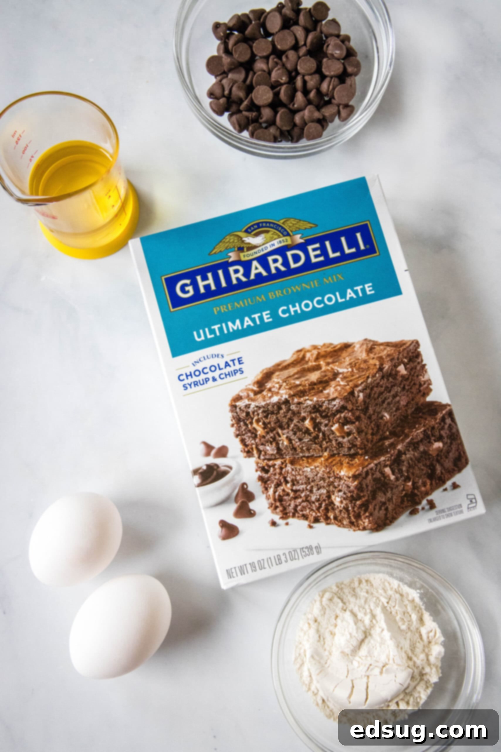 Brownie mix cookies are soft-baked, fudgy cookies made with only 4 ingredients! All you need is a box of brownie mix, oil, eggs, and flour. All the simple ingredients for making brownie mix cookies laid out on a clean white surface: a box of brownie mix, a bag of chocolate chips, a bowl of flour, vegetable oil, and two eggs.