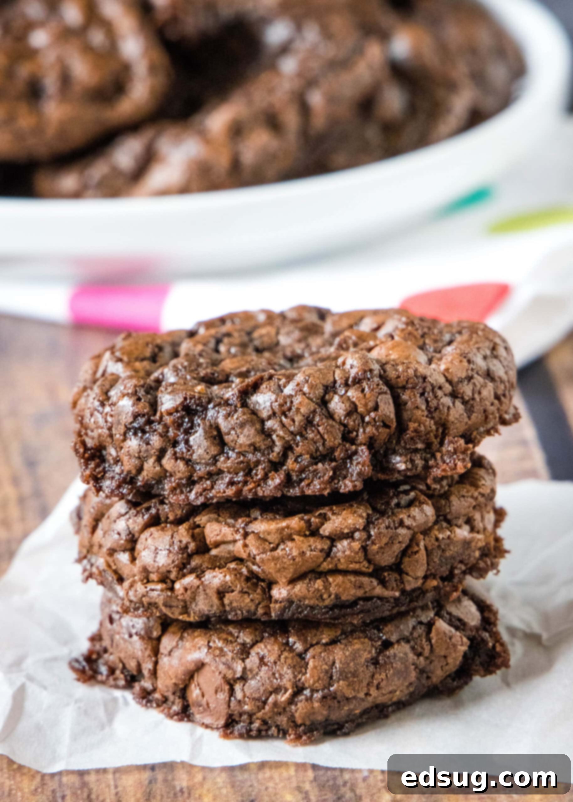 Brownie mix cookies are soft-baked, fudgy cookies made with only 4 ingredients! All you need is a box of brownie mix, oil, eggs, and flour. A beautifully arranged stack of three warm, soft-baked brownie mix cookies, gleaming with chocolate, presented on a piece of parchment paper.