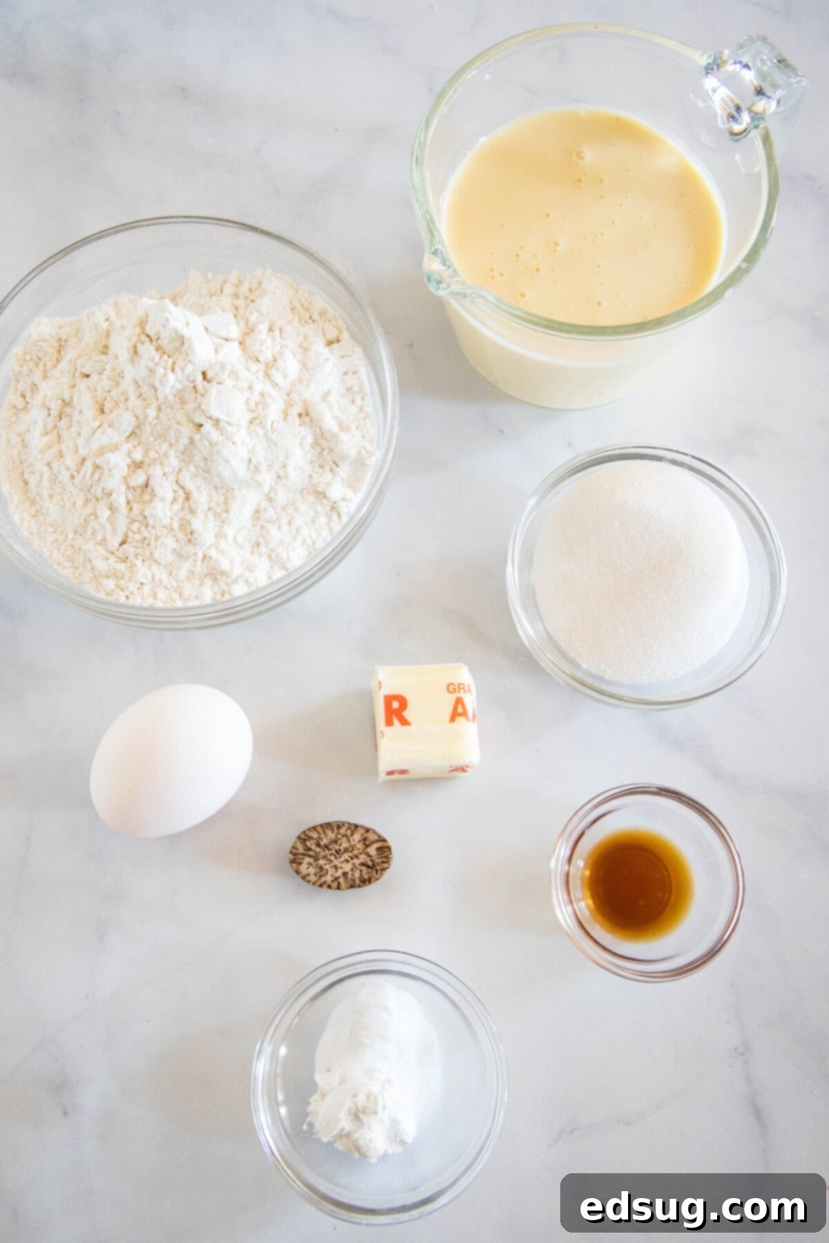 Creamy Spiced Eggnog Pancakes 3 A flat lay of eggnog pancake ingredients including flour, baking powder, baking soda, nutmeg, sugar, an egg, vanilla extract, and eggnog, neatly arranged on a kitchen surface.